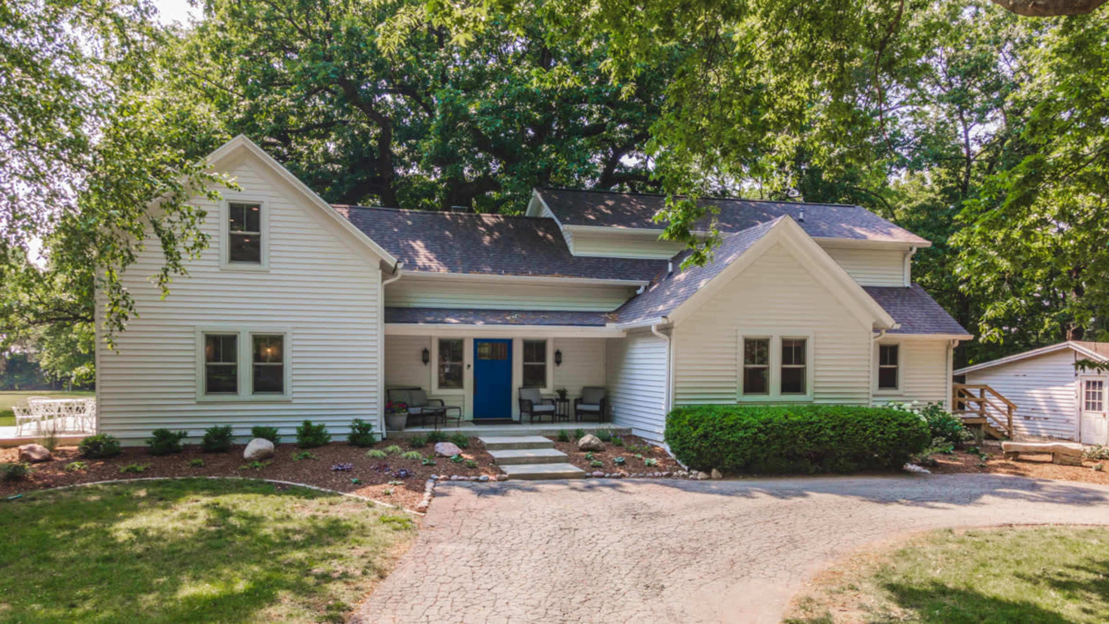 A white, two-story house with blue accents is surrounded by trees and features a front porch and a stone pathway leading to the entrance.