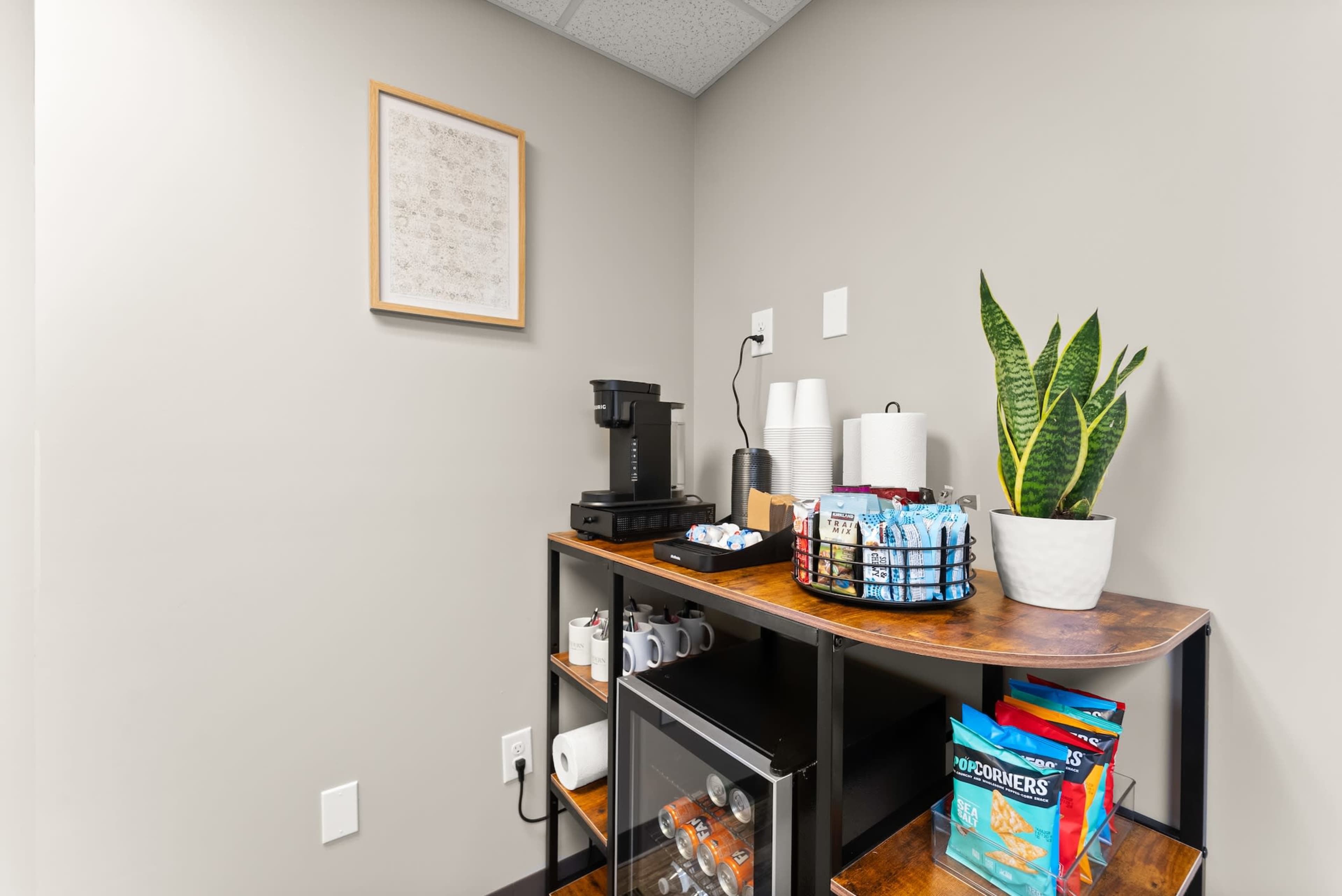 A coffee station is arranged in a corner, featuring a coffee maker, cups, a plant, and snacks displayed on a wooden shelf.