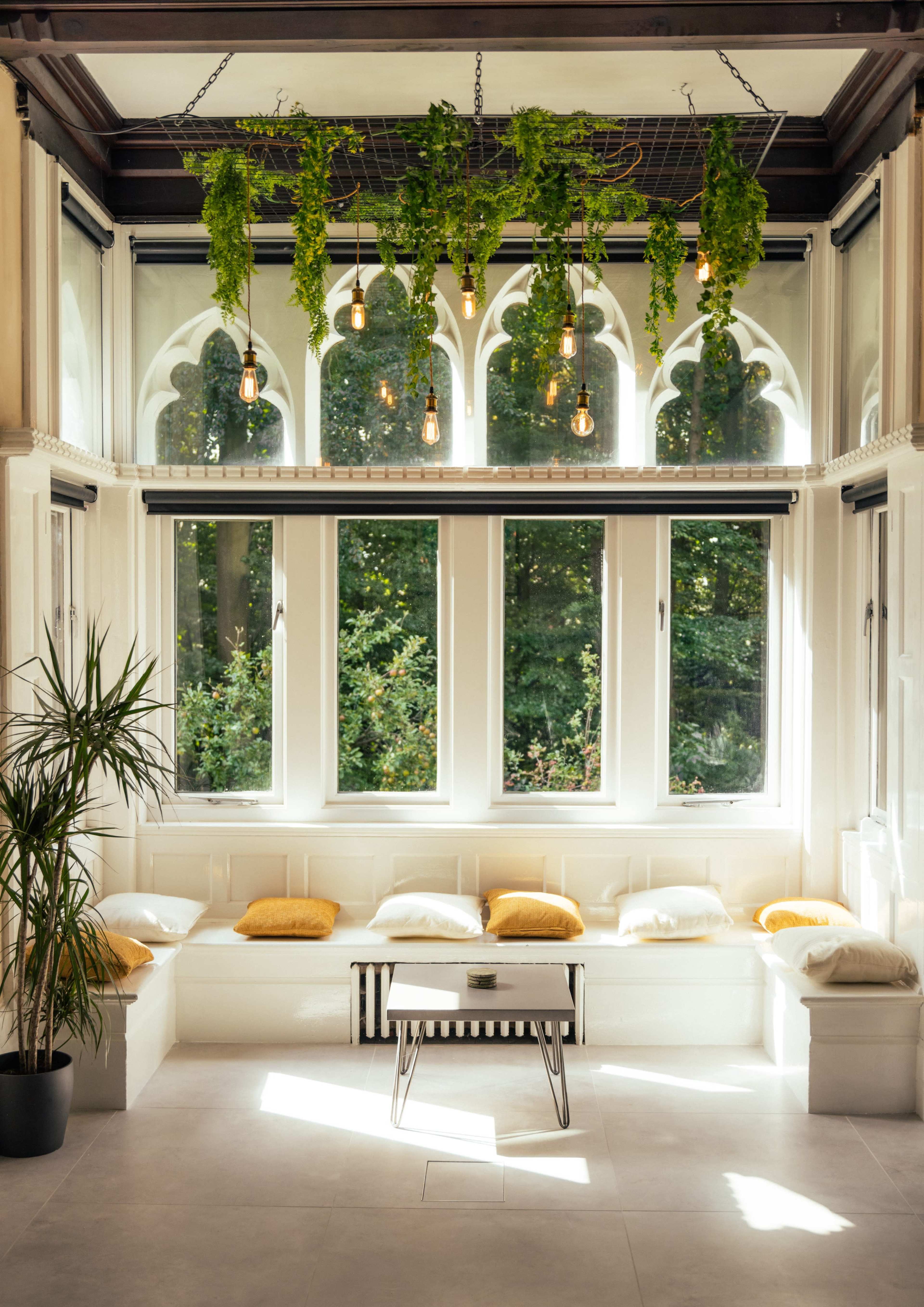 A spacious indoor seating area features large windows, hanging plants, and a central table surrounded by cushions.