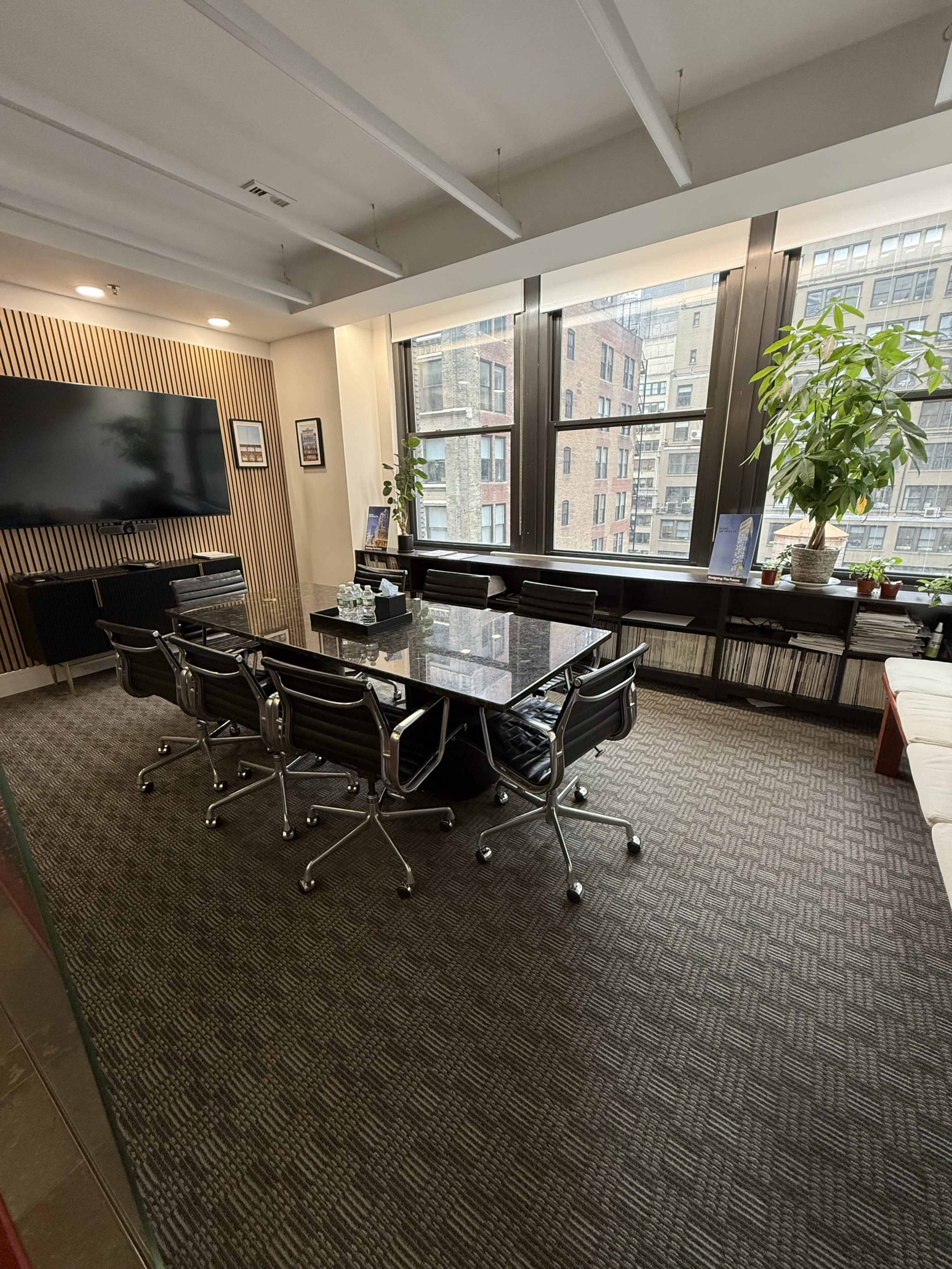 The image shows a modern conference room with a large table surrounded by black chairs, a wall-mounted television, and large windows displaying a cityscape.