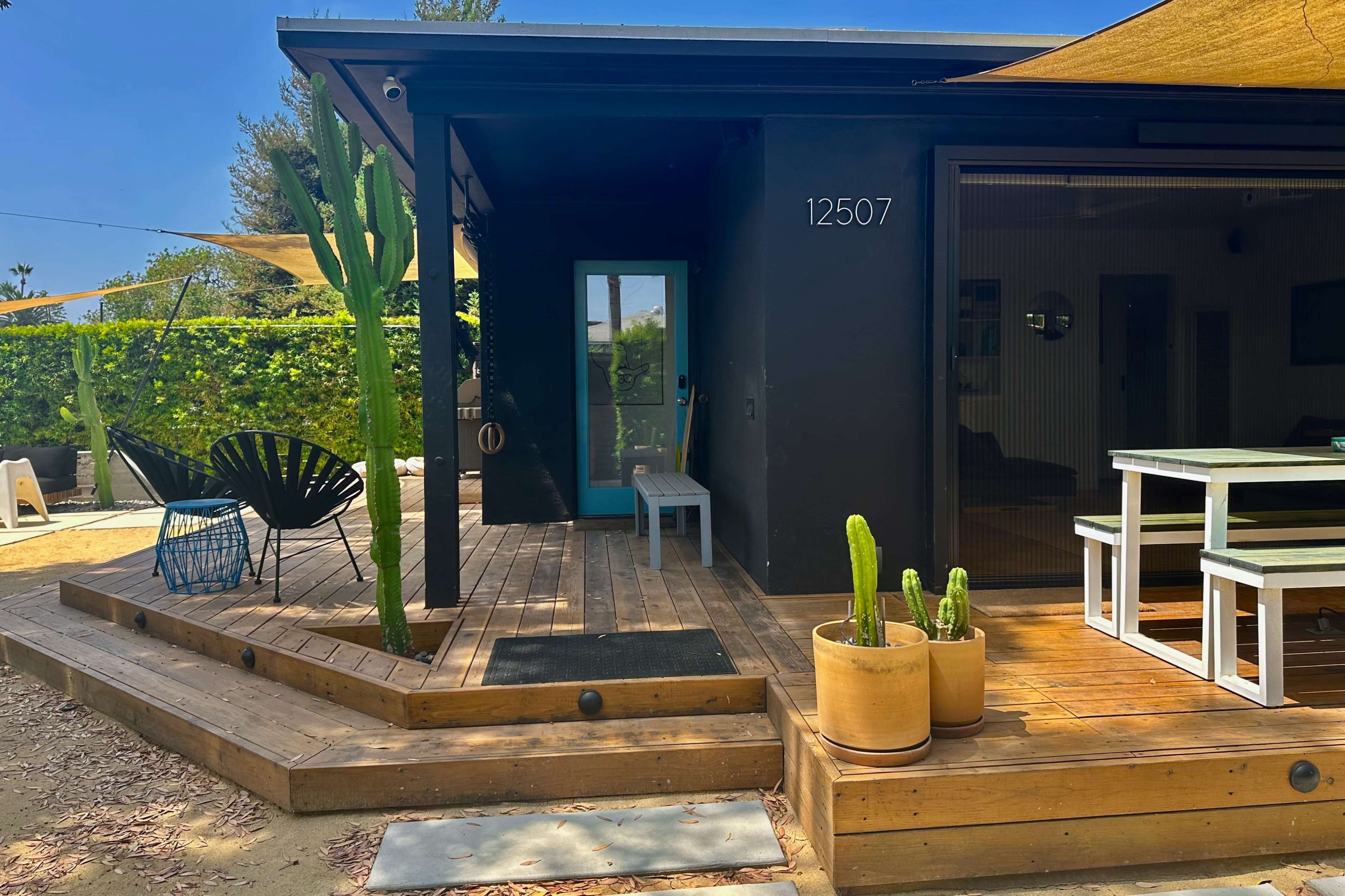 A modern wooden deck with cacti and a small table leads to a black house featuring a blue door.