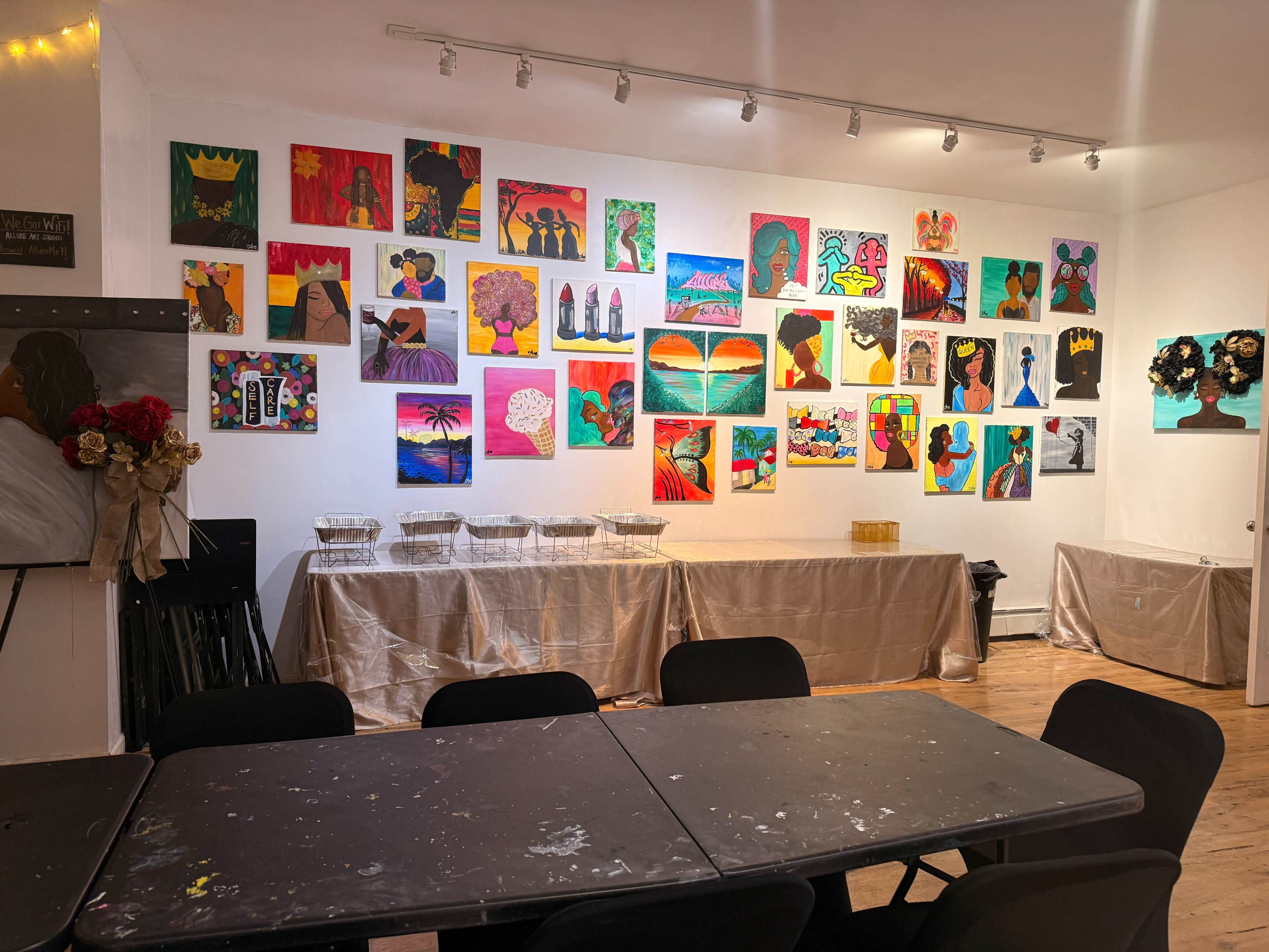 The image shows a room with a table in the foreground and a wall adorned with various colorful paintings.