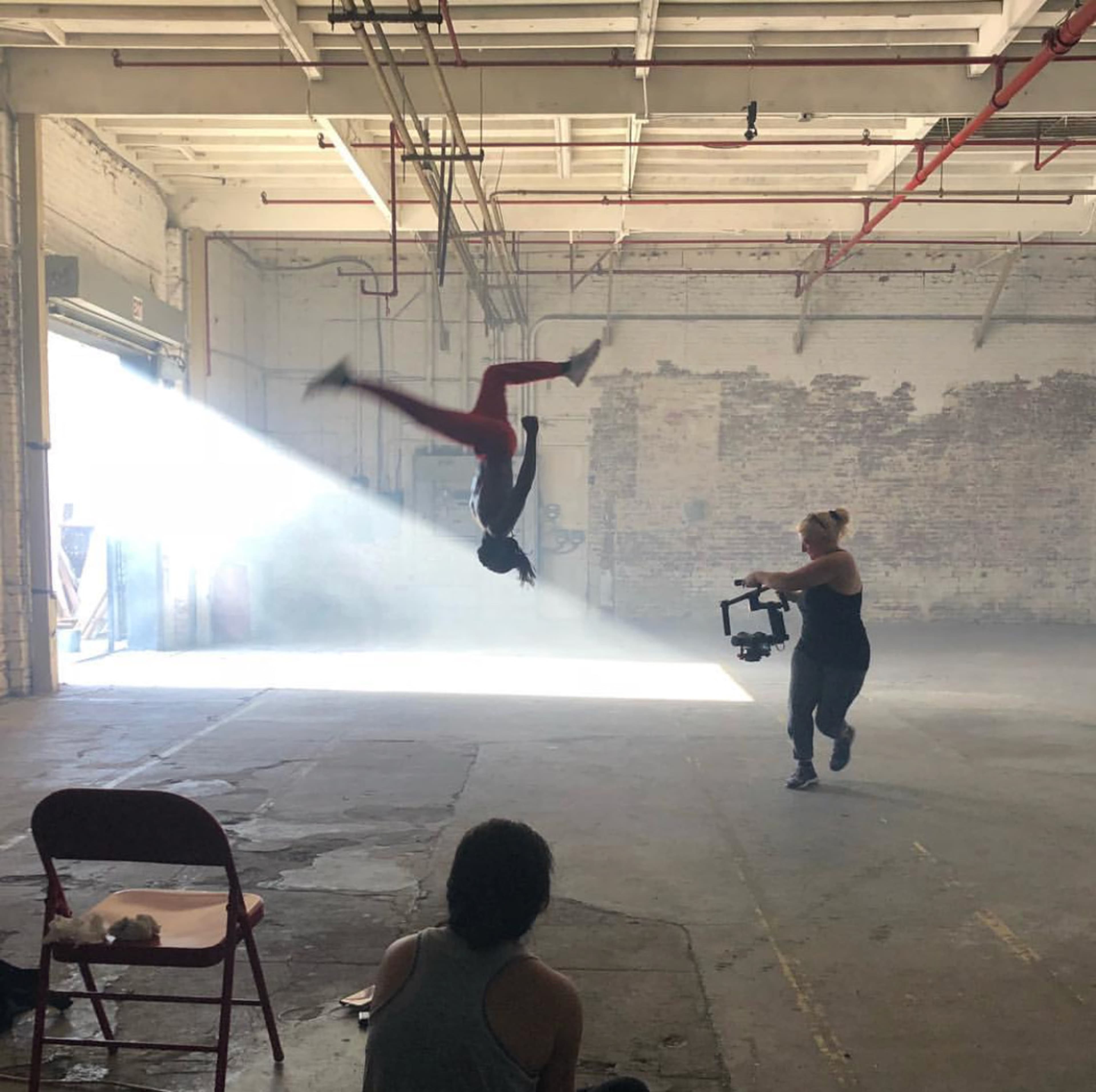A person performs an acrobatic flip in an industrial space while another individual captures the moment with a camera.