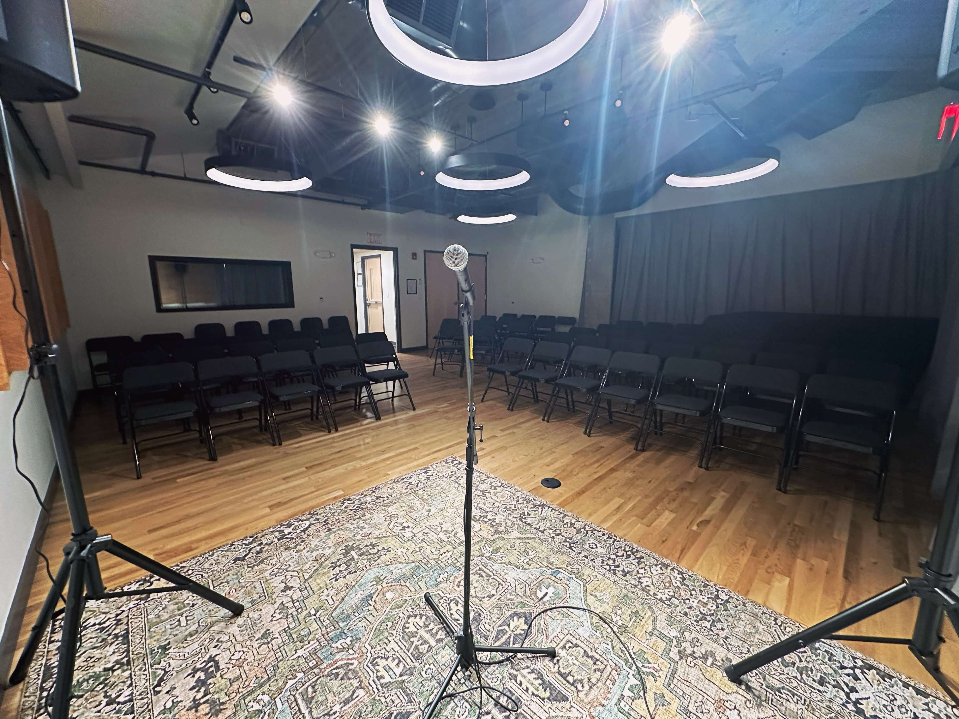 A microphone stands on a stand in front of a row of empty chairs arranged in a well-lit room with a patterned rug.