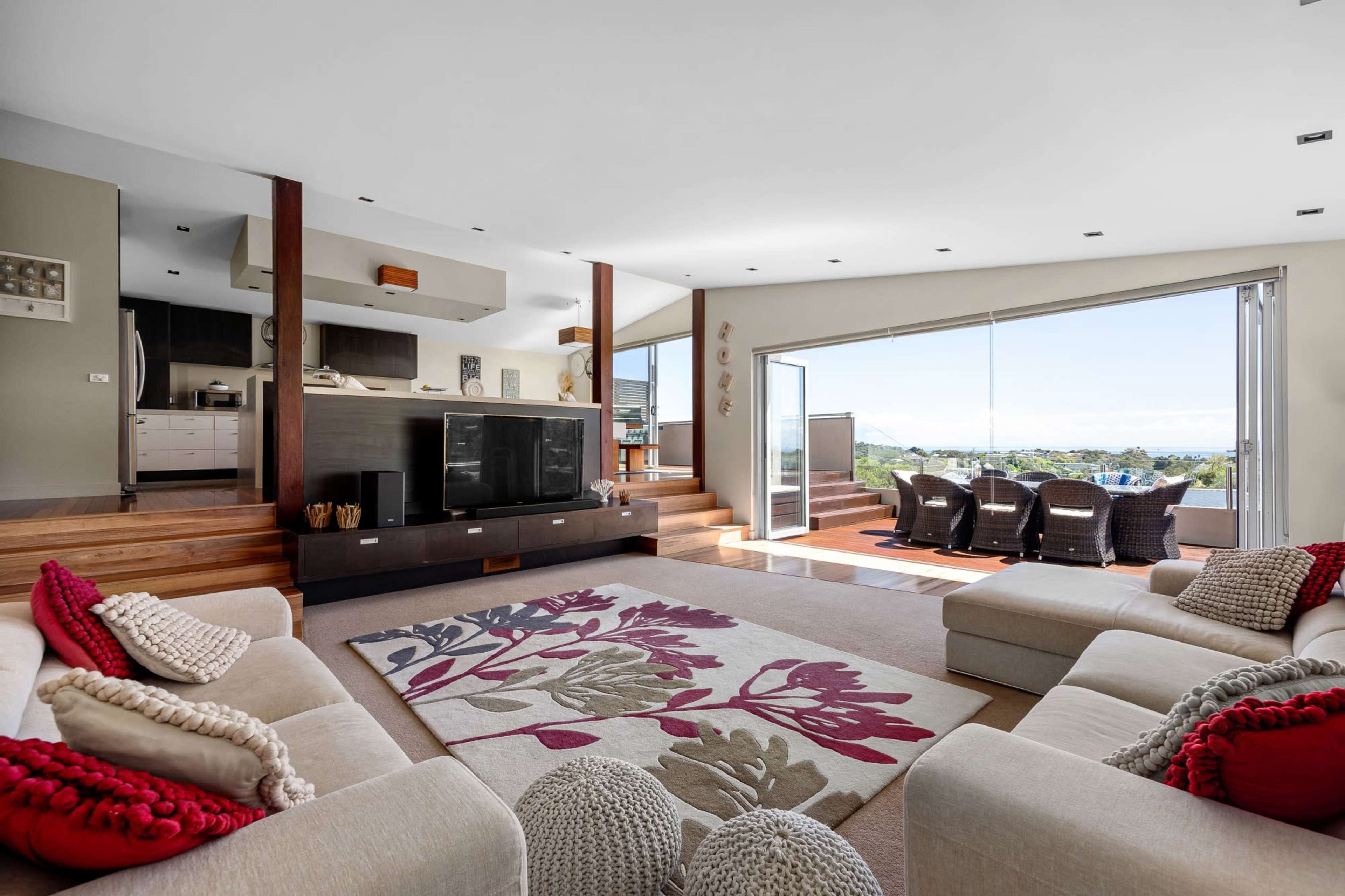 The living room features a spacious layout with a large area rug, two sofas, a modern kitchen in the background, and large sliding glass doors that open to a view outside.