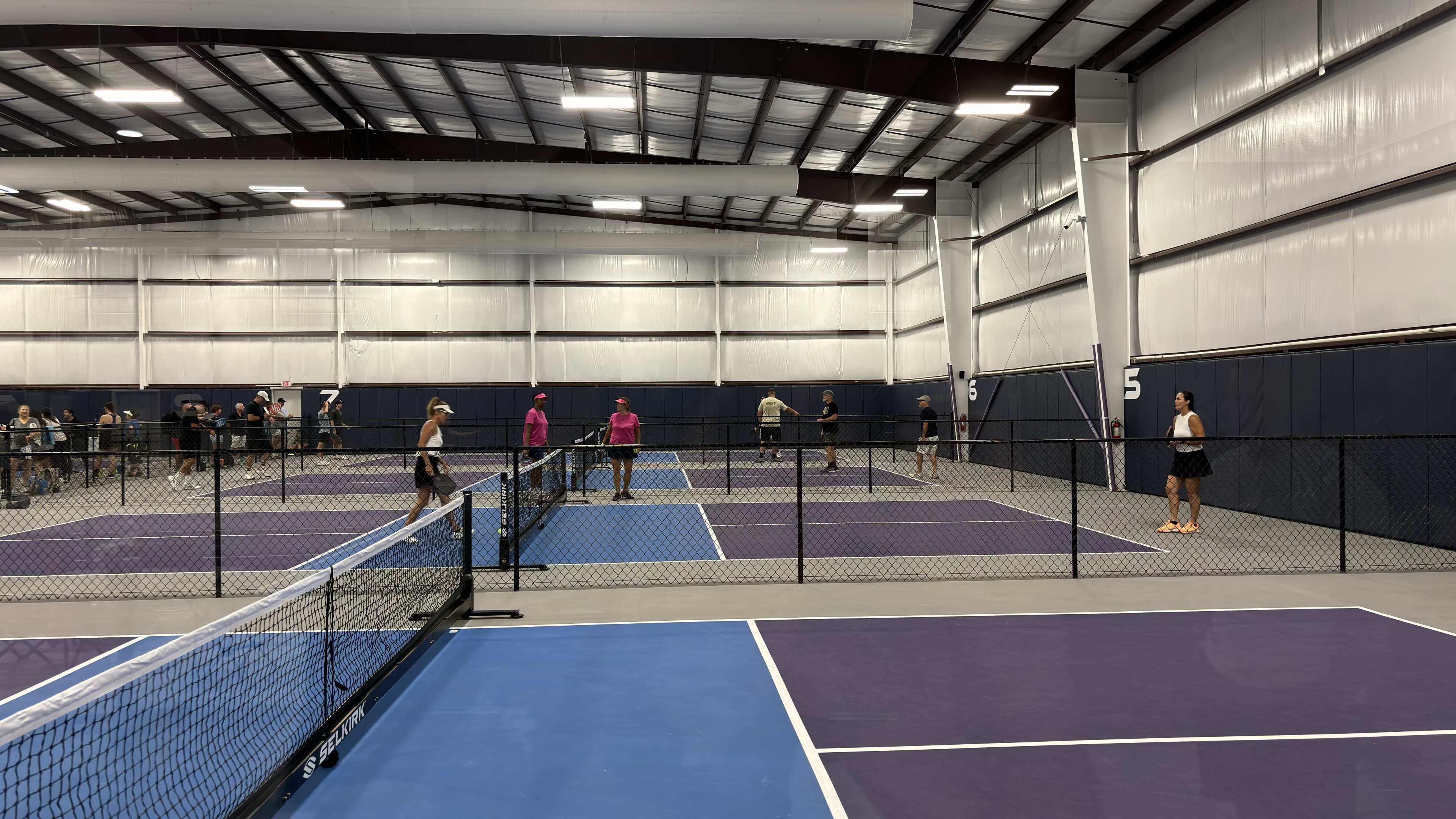 The image shows an indoor tennis facility with multiple courts, where several players are engaged in matches while others observe from the sidelines.