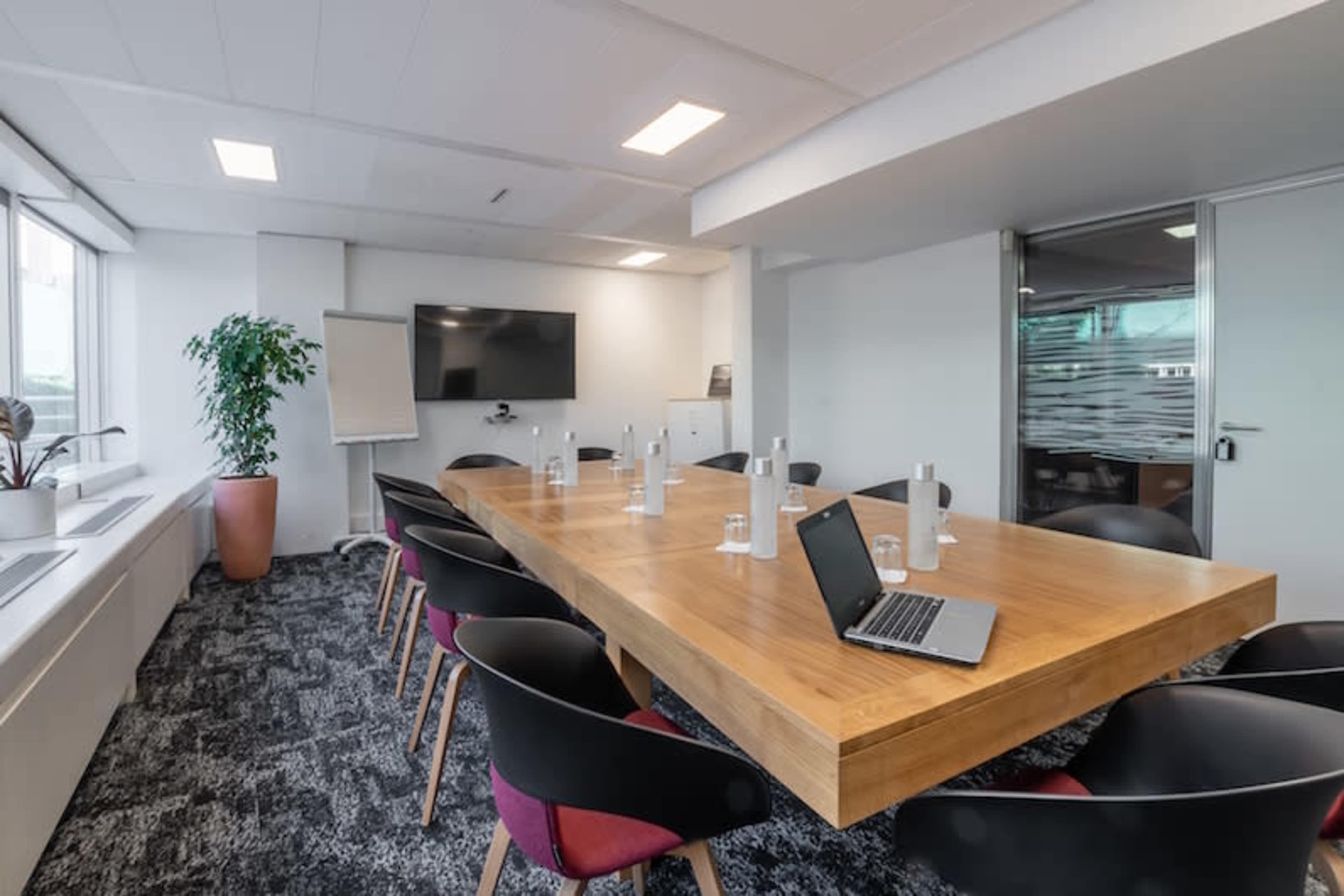 A modern conference room features a large wooden table surrounded by black chairs, with a laptop open on the table and a television screen mounted on the wall.