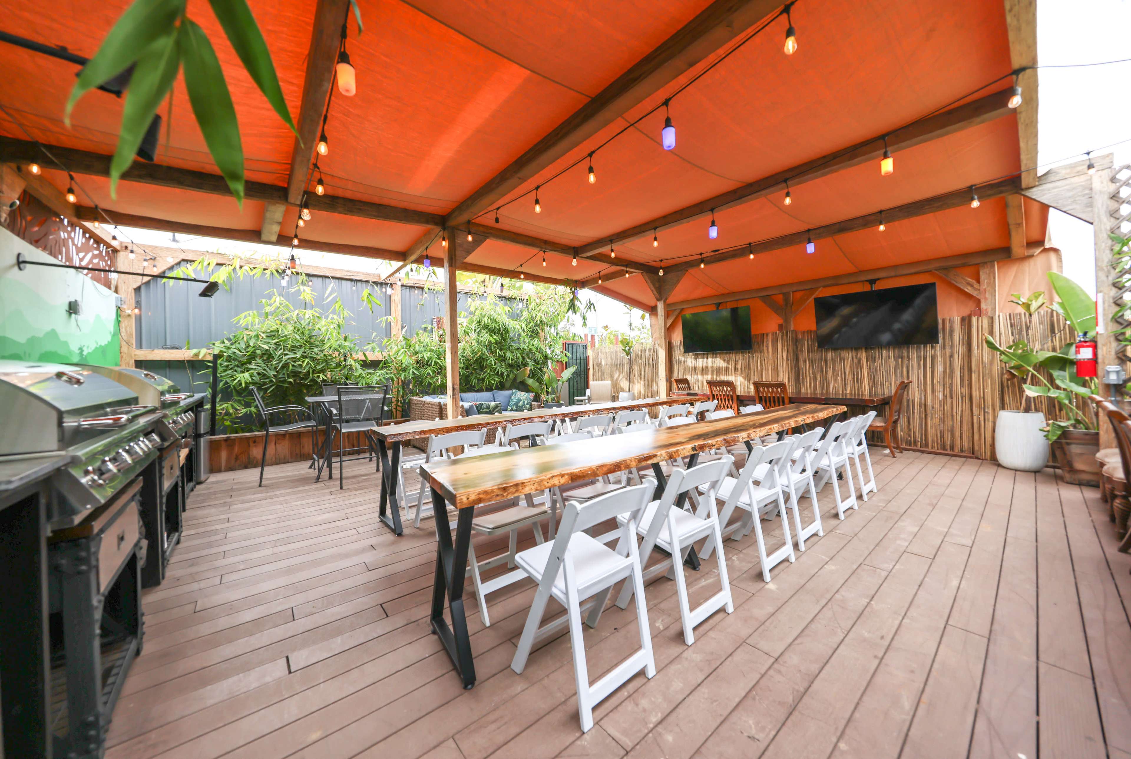 An outdoor dining area with several long tables underneath an orange canopy, surrounded by greenery and featuring string lights.
