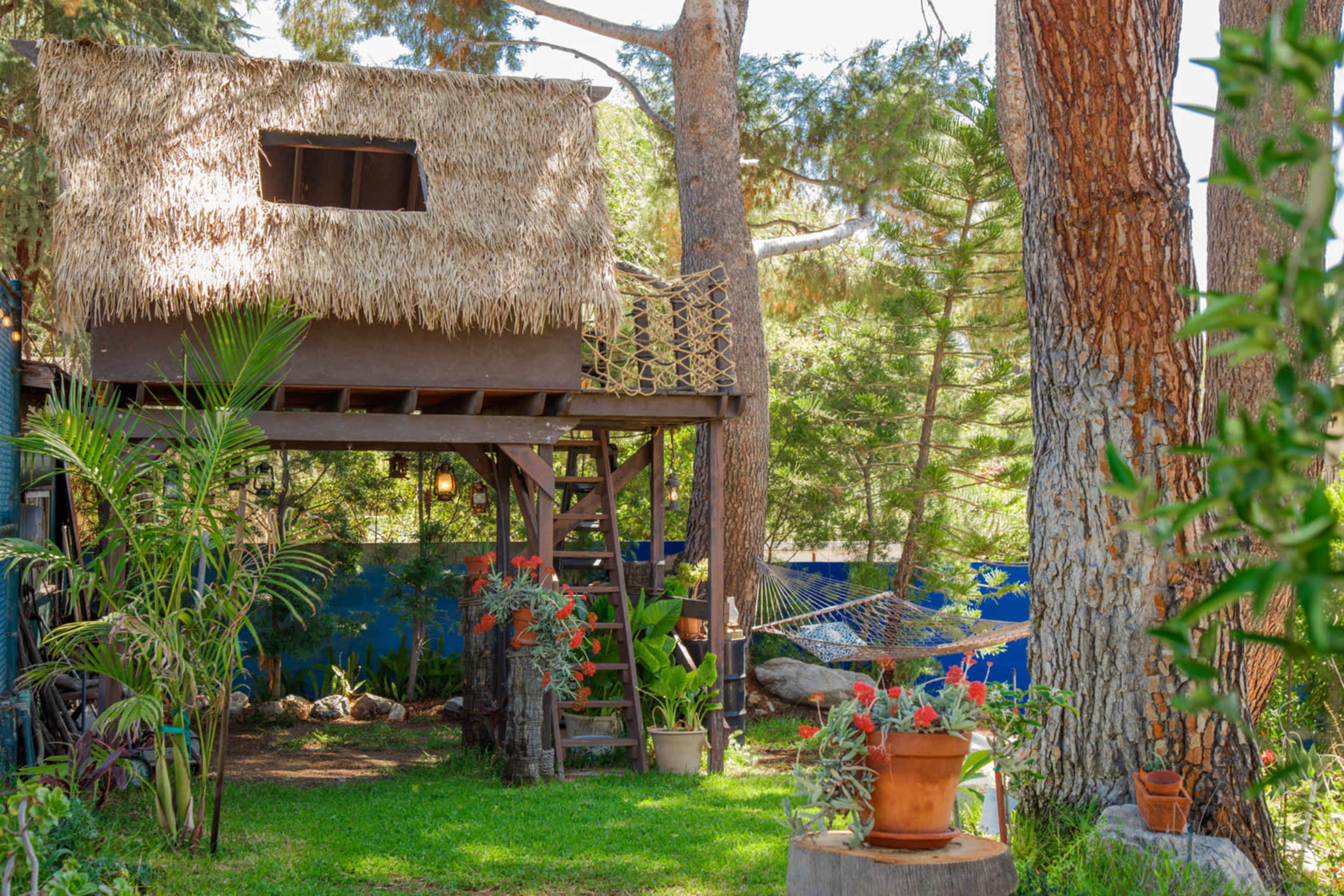 A thatched-roof treehouse is perched among tall trees in a garden filled with colorful plants and a hammock nearby.