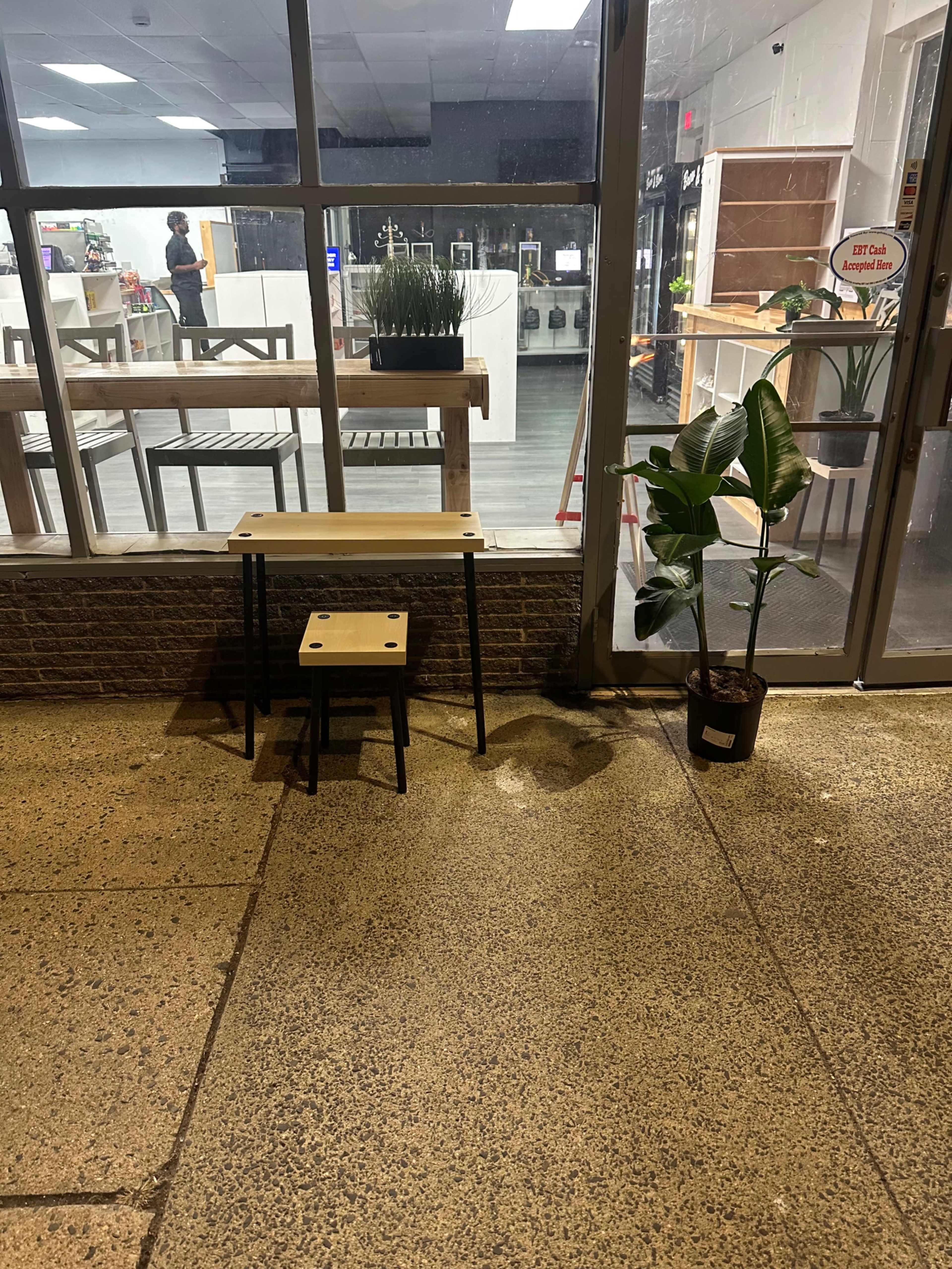 The image shows a small wooden table and stool positioned outside a shop, alongside a potted plant, with a view of the interior behind large glass windows.