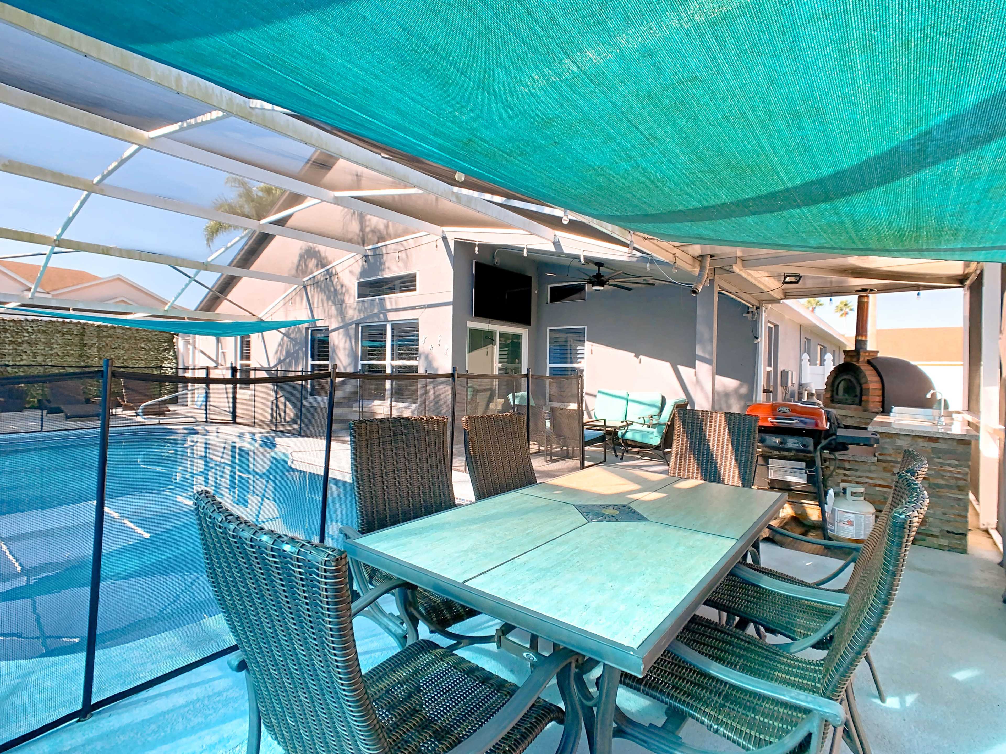 A patio area features a table with several chairs beside a swimming pool, shaded by a tarp and surrounded by a screened enclosure.