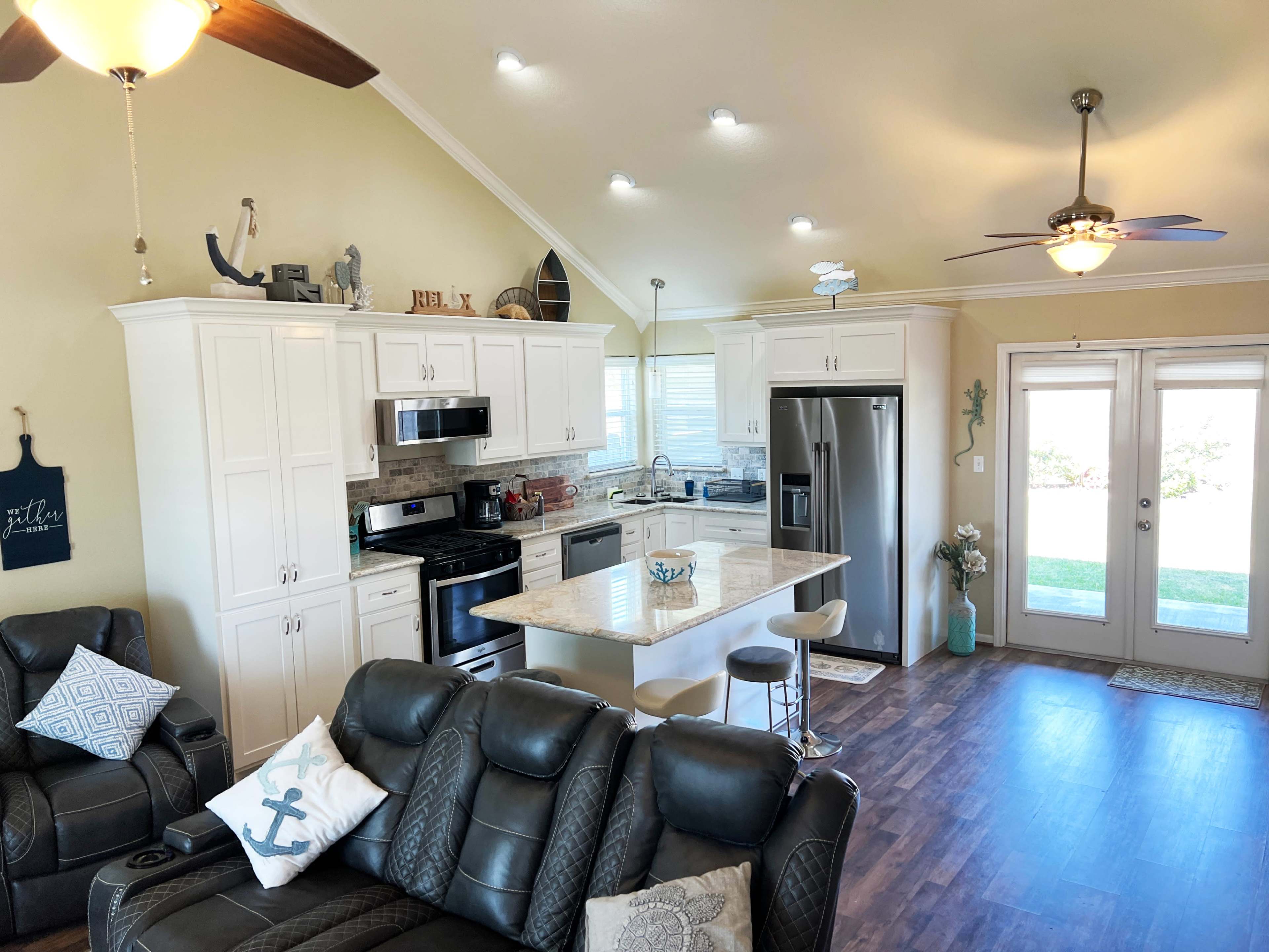 A modern kitchen with white cabinetry, a large island, and stainless steel appliances connects to a living area featuring dark leather furniture.