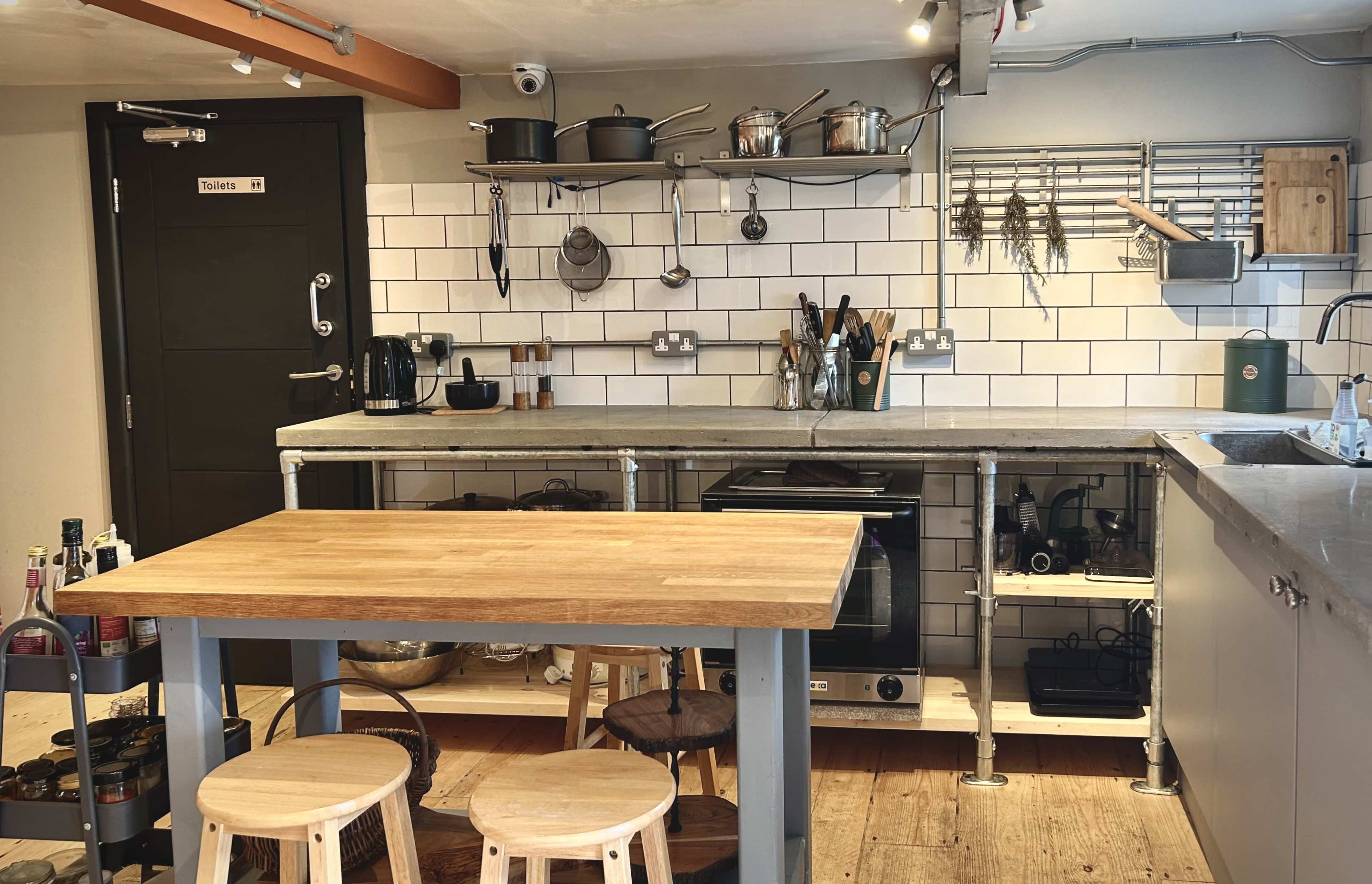 The image shows a modern kitchen with a wooden table, metal stools, concrete countertops, and various cooking utensils hanging on the wall.