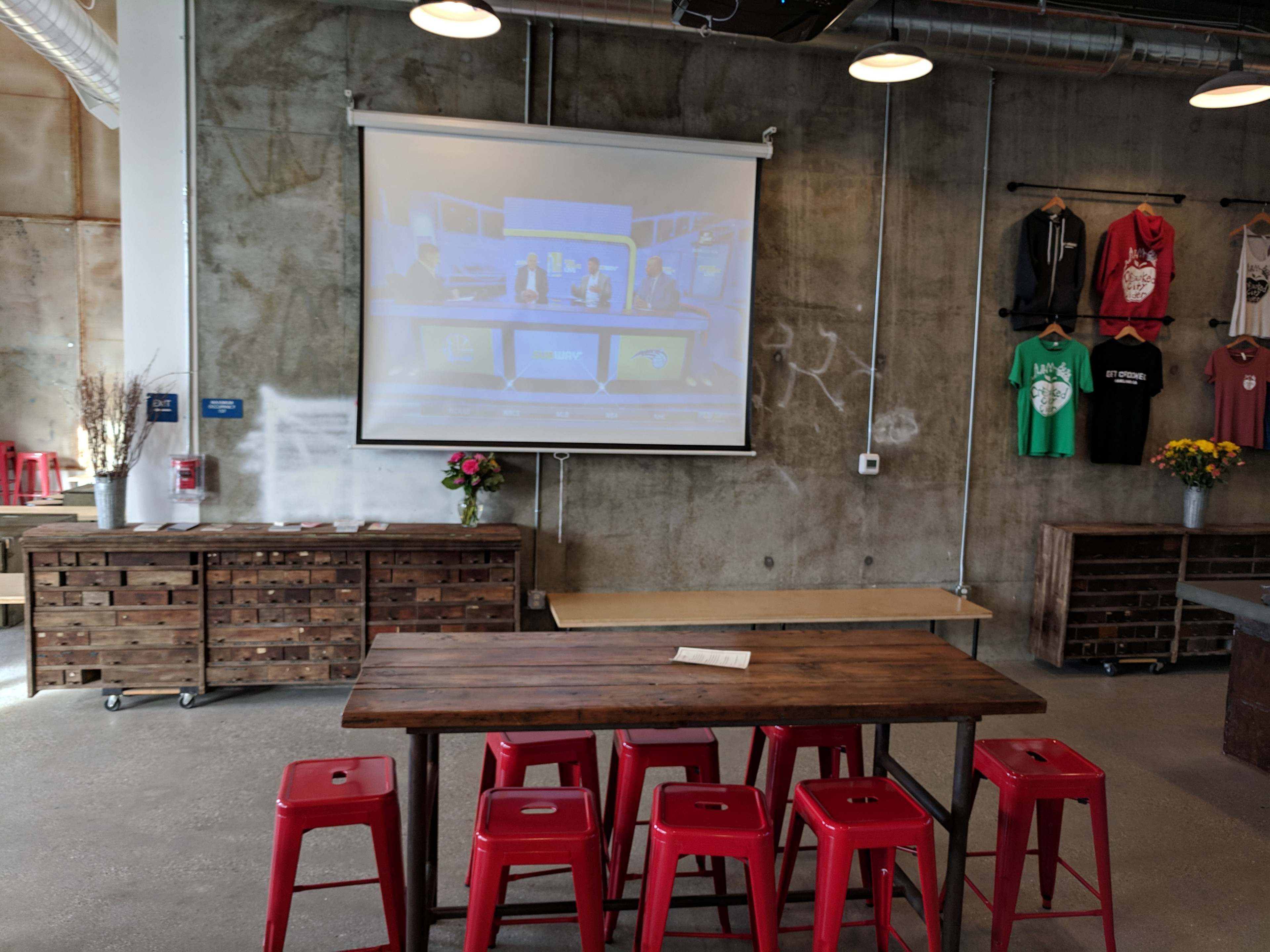 A wooden table with red stools is positioned in front of a projection screen displaying a video in a modern, industrial-style room.