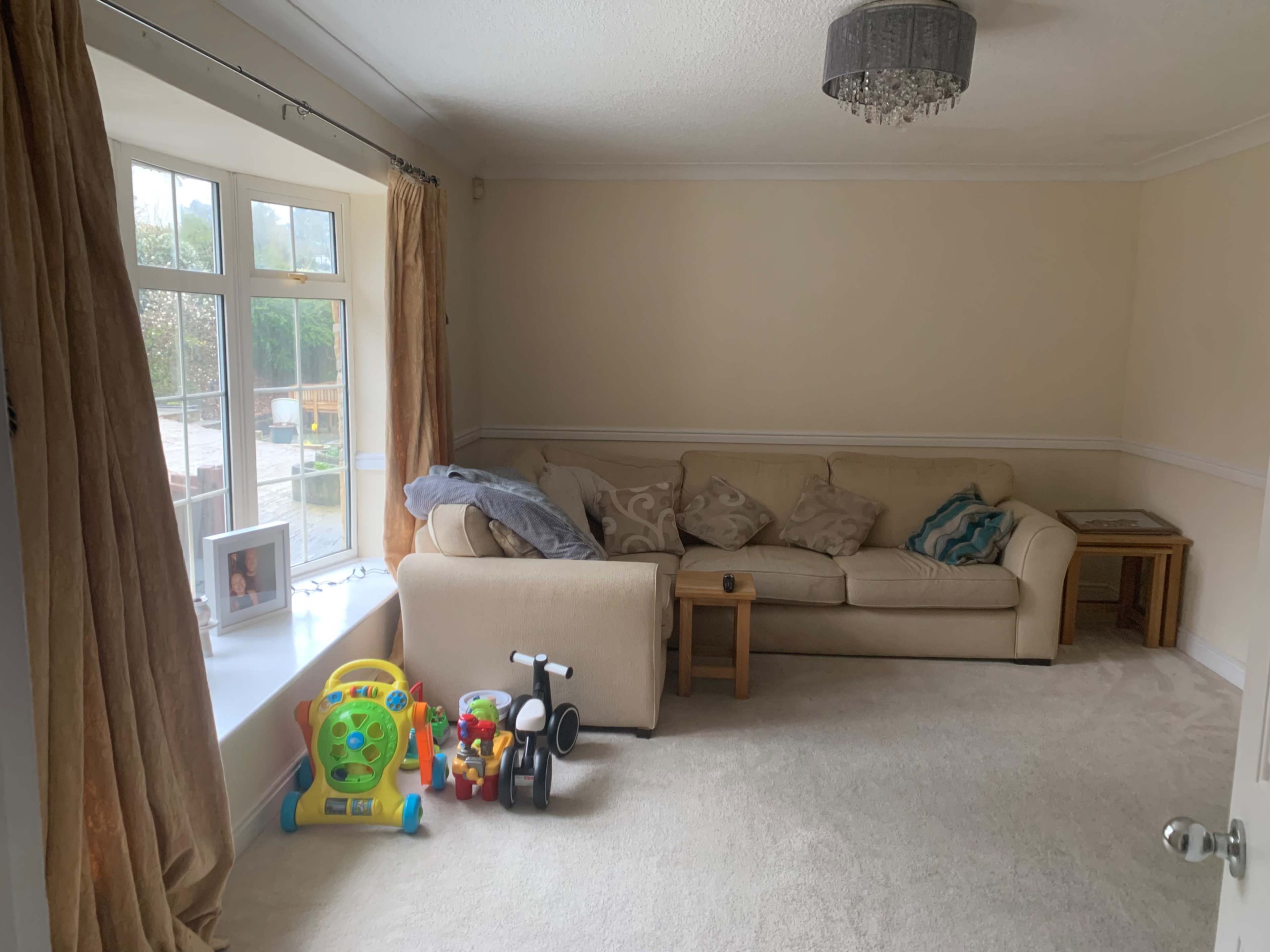 The image shows a light-colored living room with a beige couch, a small coffee table, and children's toys near a large window.