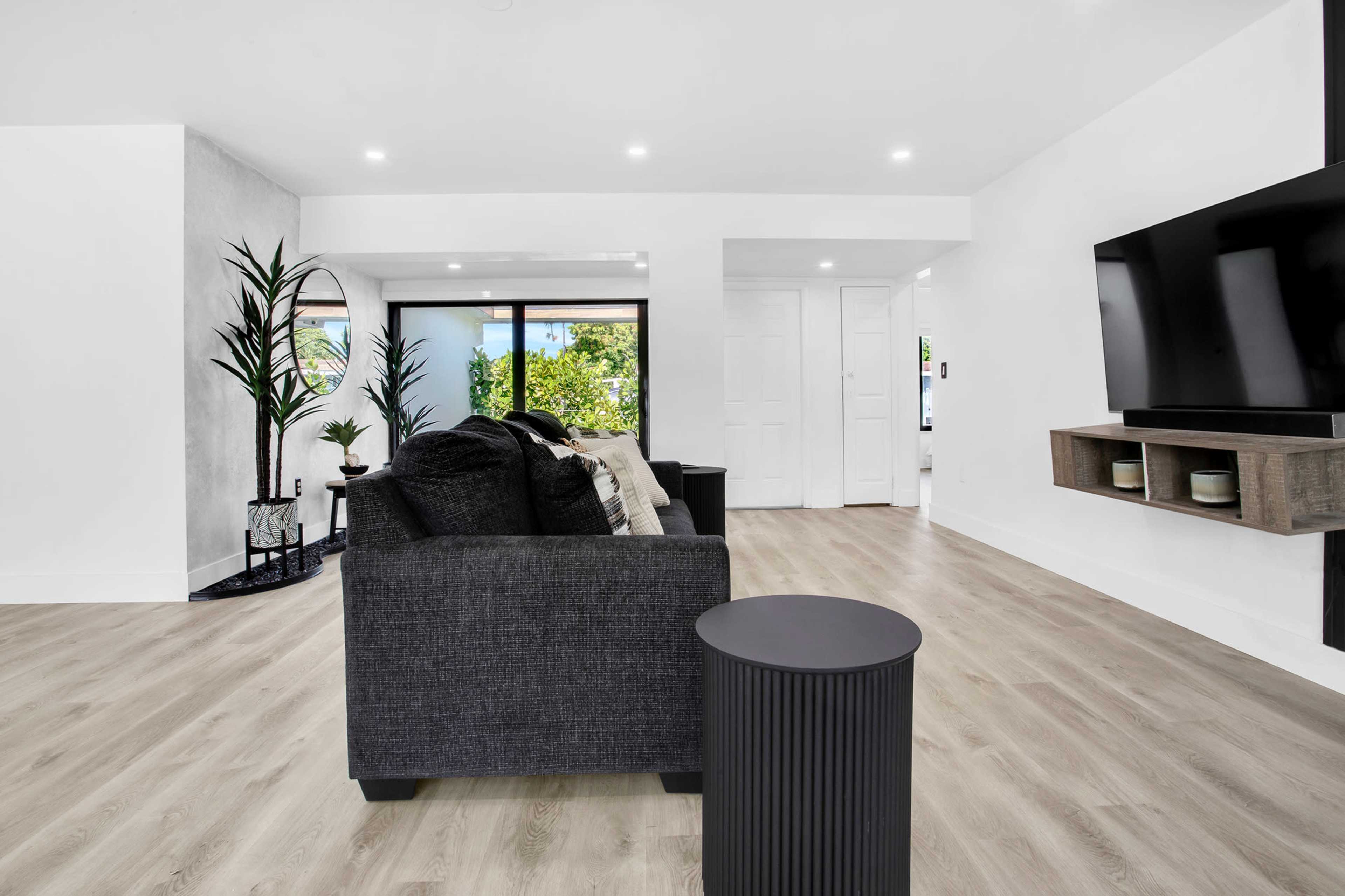 The image shows a modern living room with a dark sofa, a small side table, and large windows allowing natural light to enter.