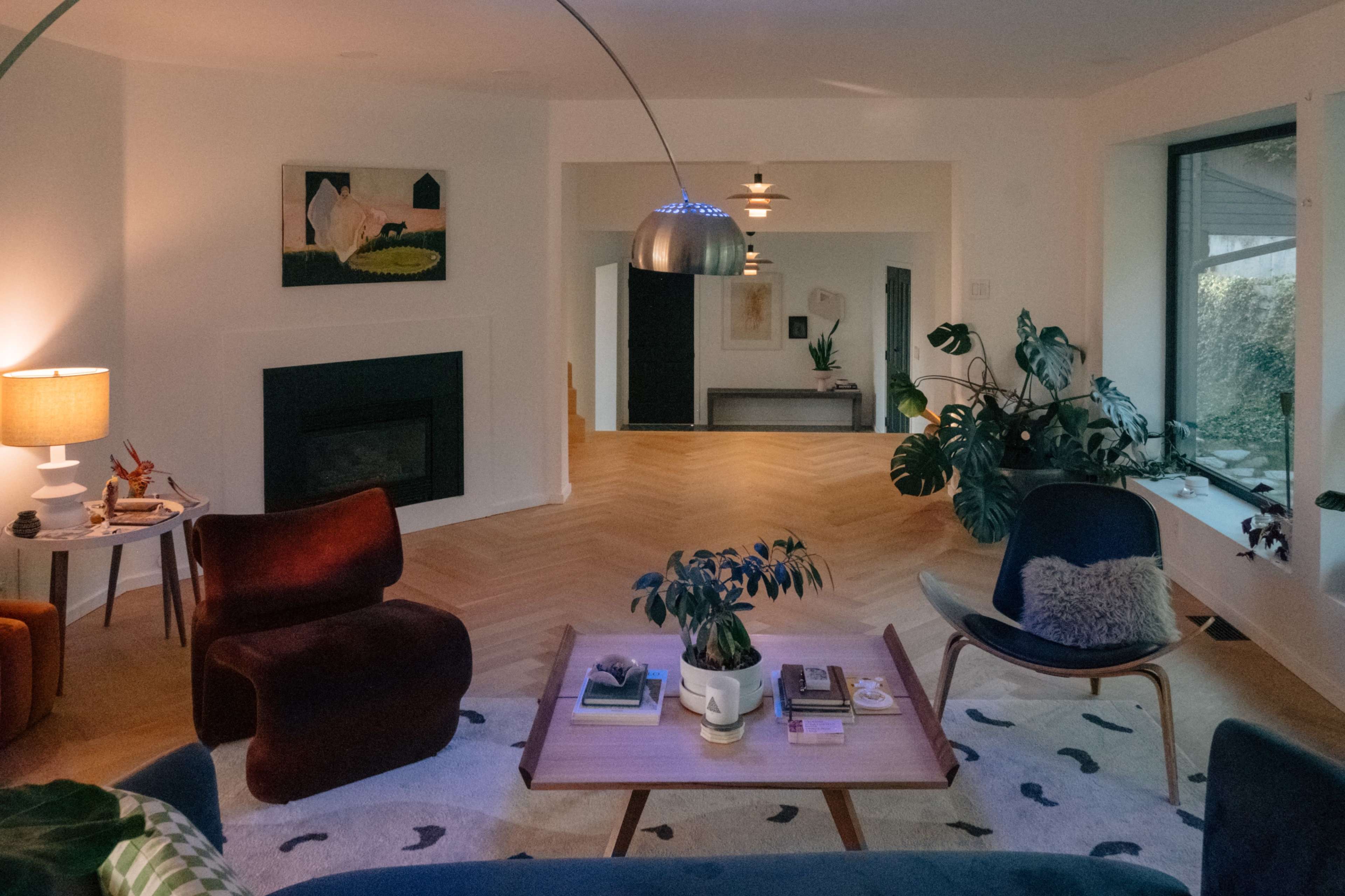 The image shows a modern living room with a fireplace, various seating arrangements, a coffee table with decor, and large windows letting in natural light.