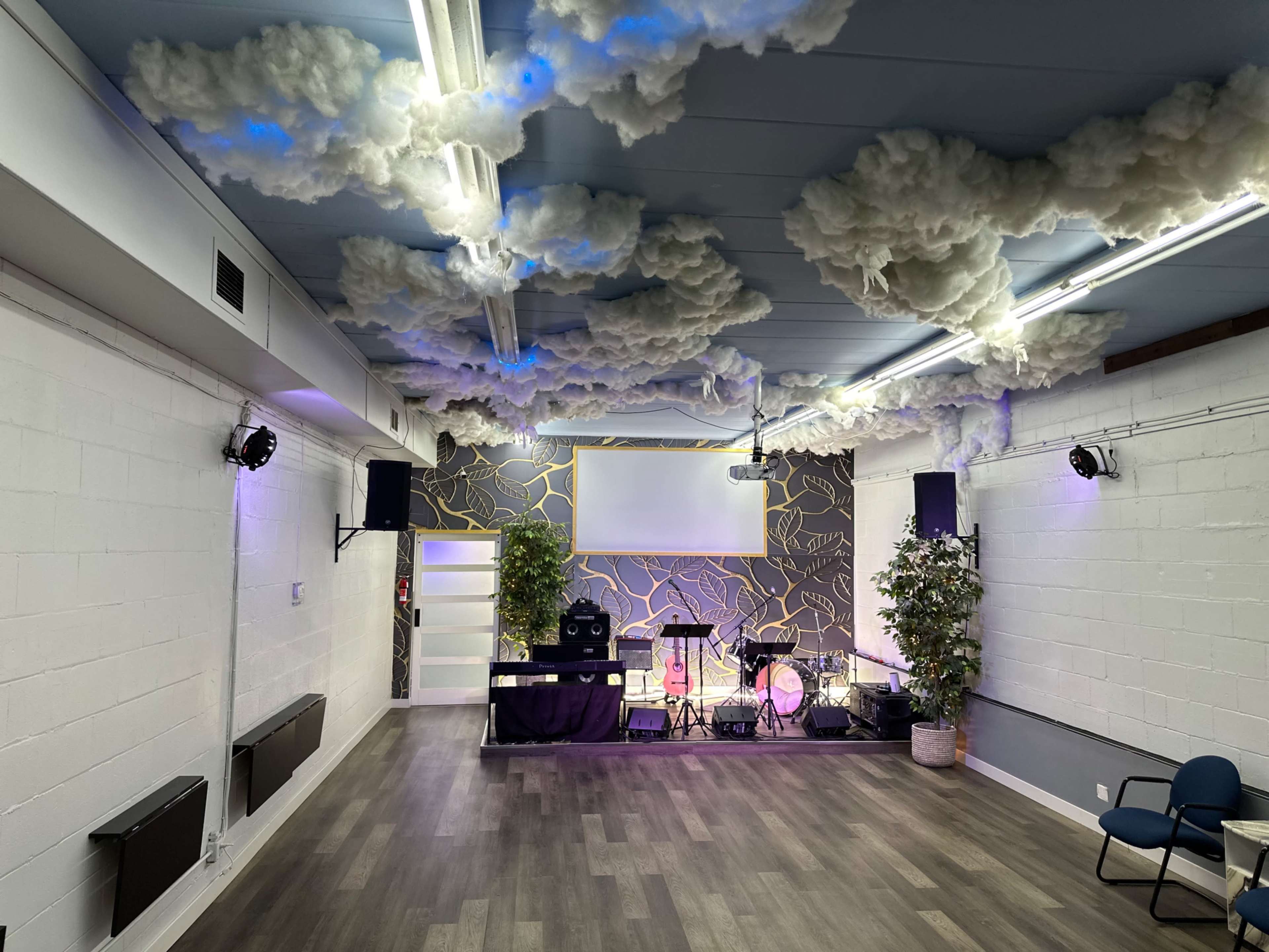 The image shows an interior space designed for events, featuring a stage with musical instruments, cloud-like decor on the ceiling, and plants positioned around the room.