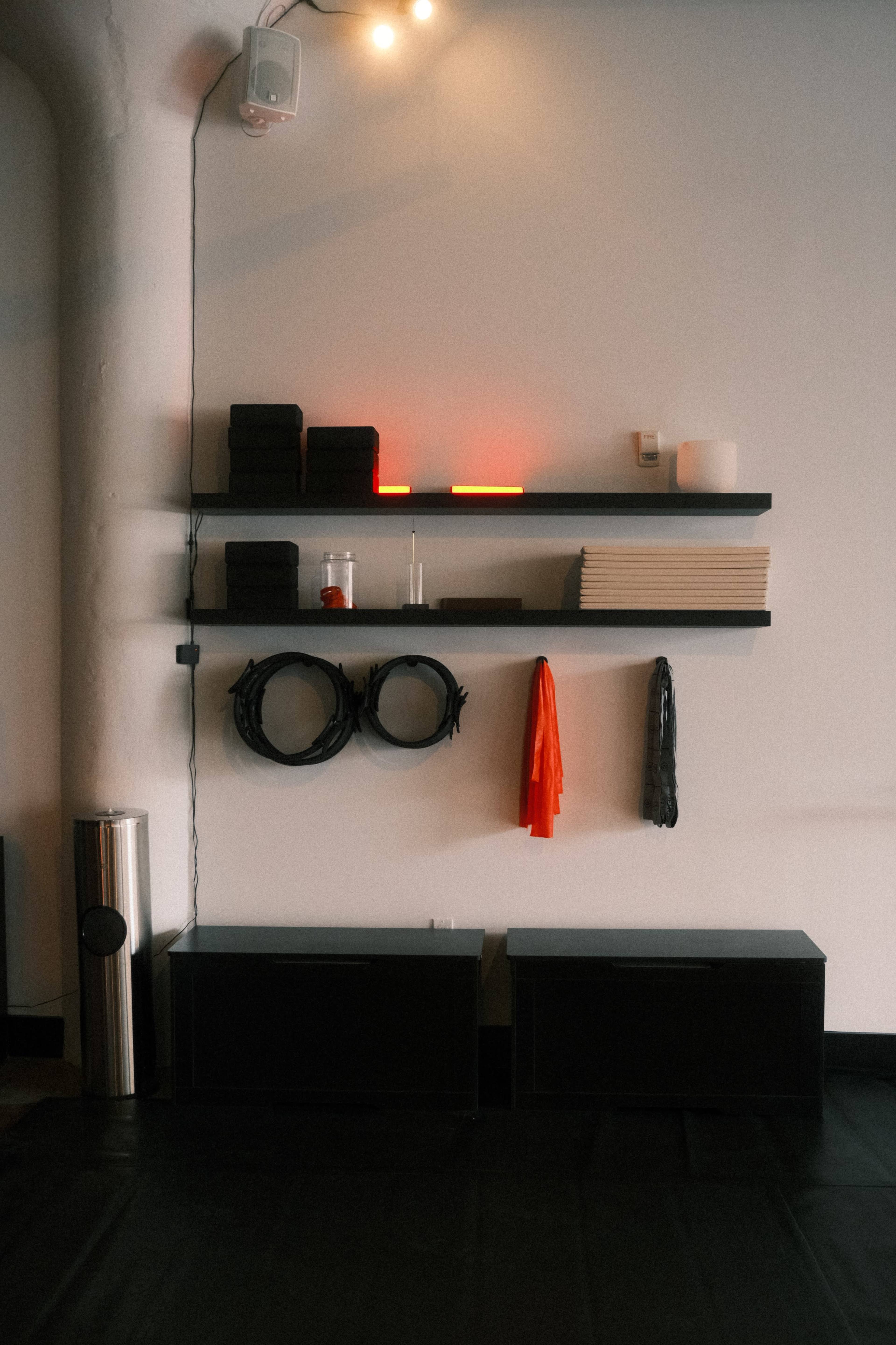 The image shows a minimalist interior wall with black shelves displaying stacked items and a few accessories, accompanied by a sleek, cylindrical object below.