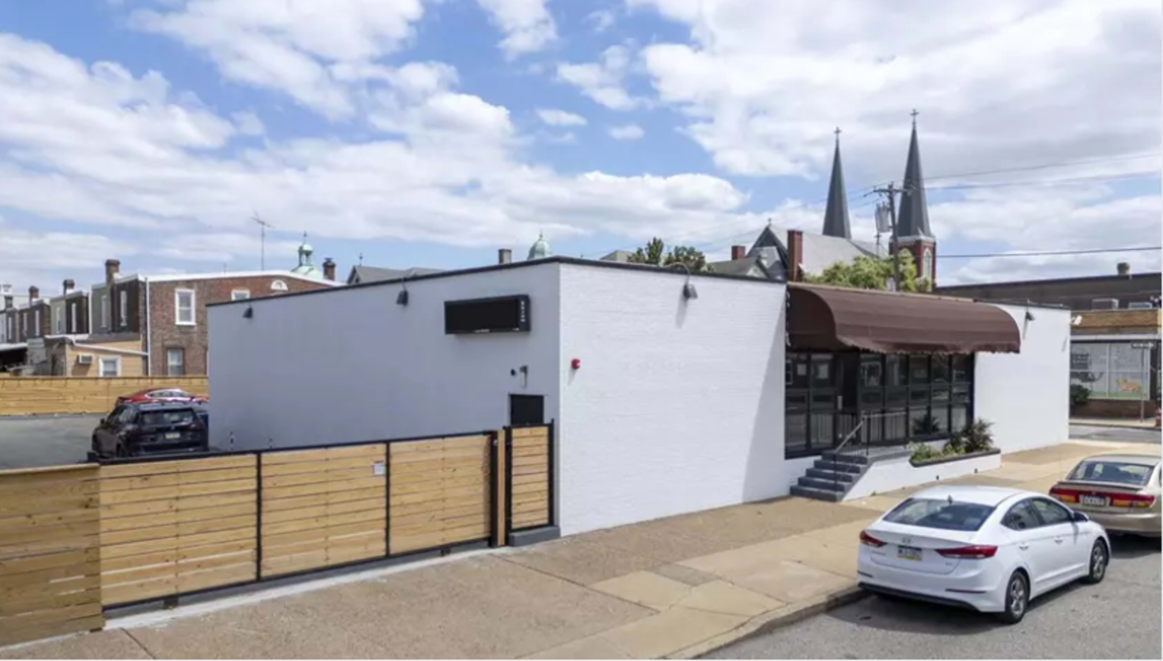 The image shows a modern, white building with a brown awning, surrounded by a wooden fence and parked cars, set against a backdrop of a cloudy sky and church spires.
