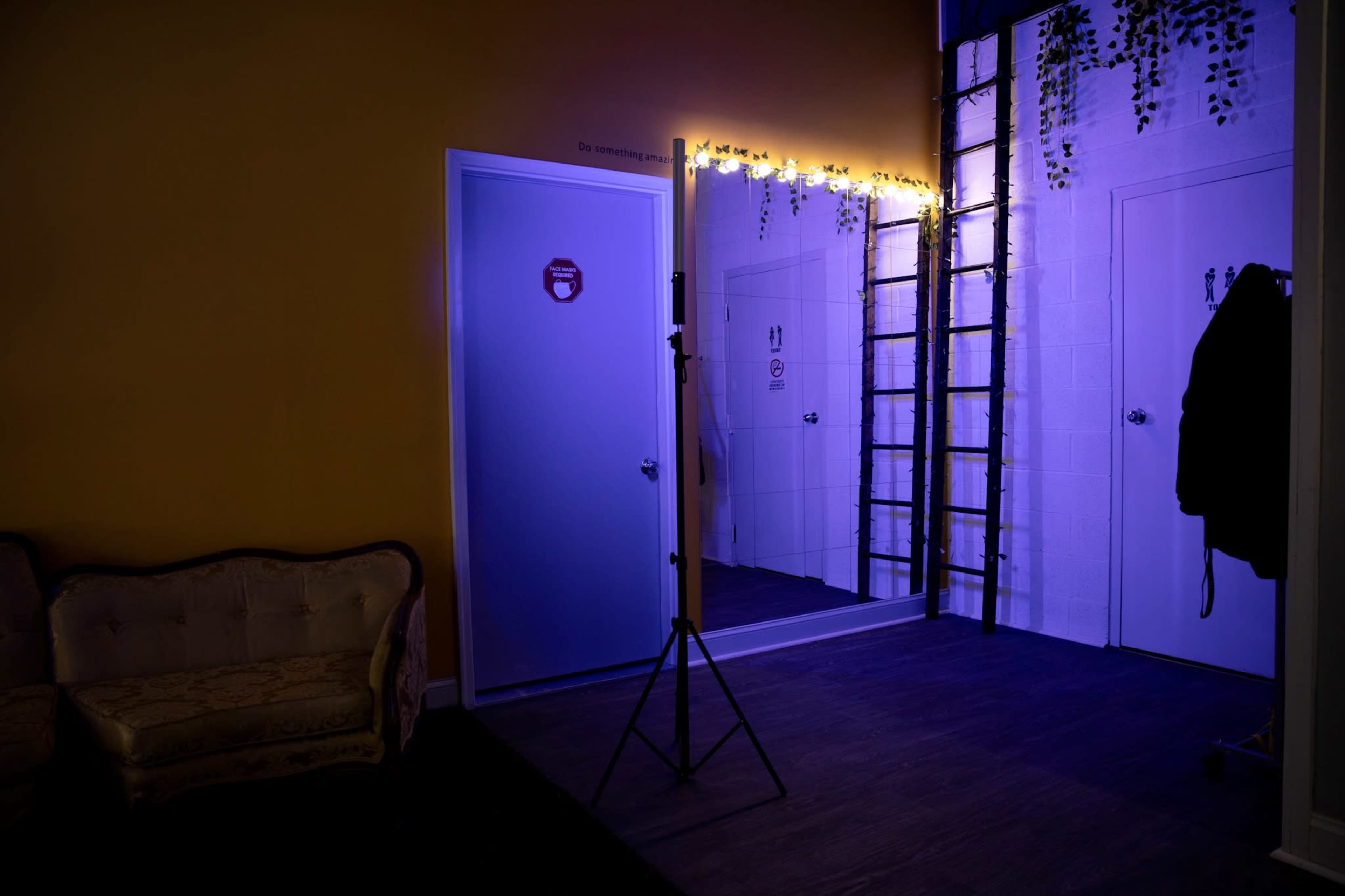 A room features a ladder against a wall, a large mirror, and illuminated signage for restrooms, with a yellow backdrop and a couch on one side.