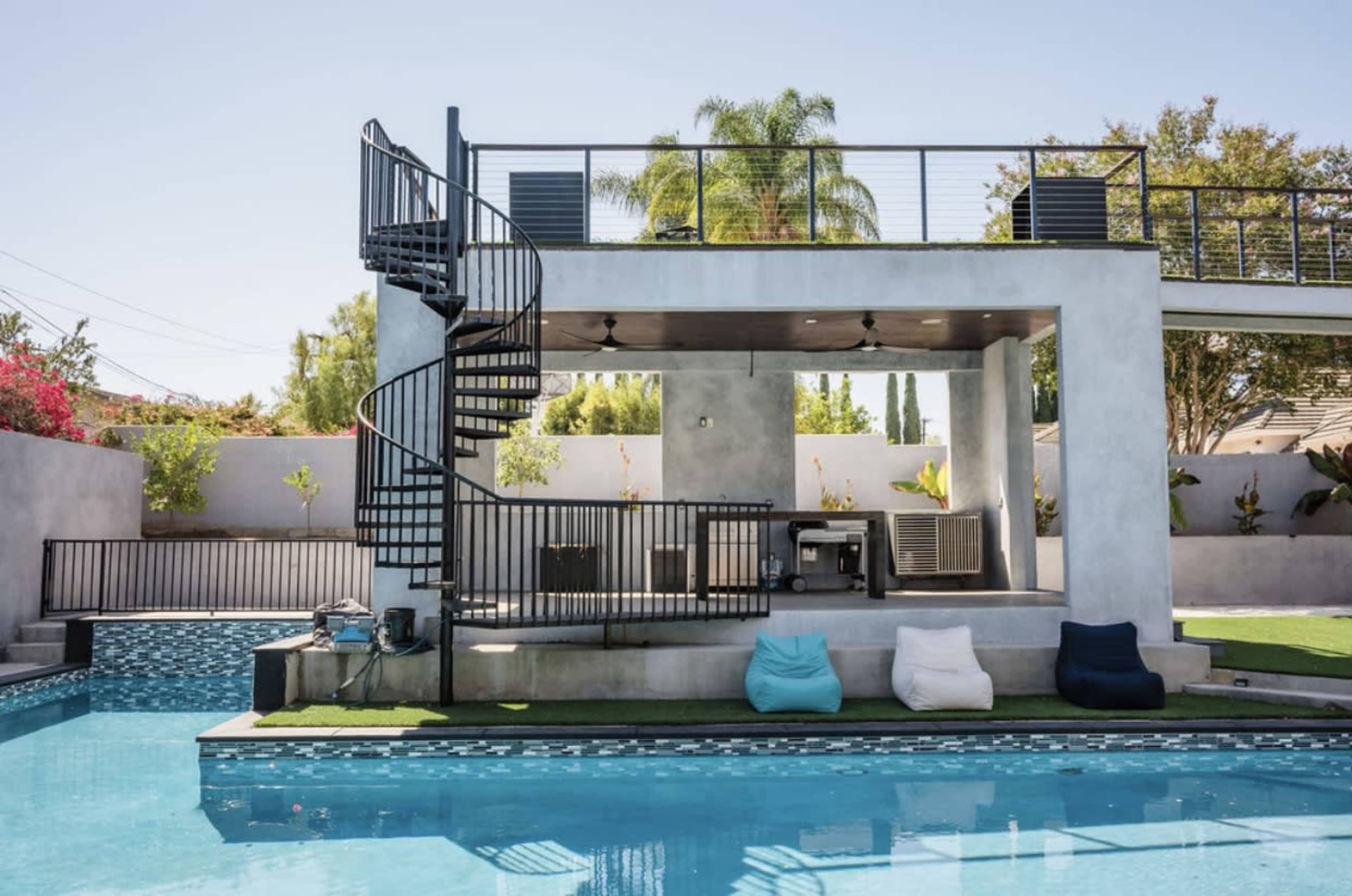 A modern outdoor space features a spiral staircase leading to a covered patio with an outdoor kitchen, alongside a swimming pool with geometric tile designs and bean bag seating.