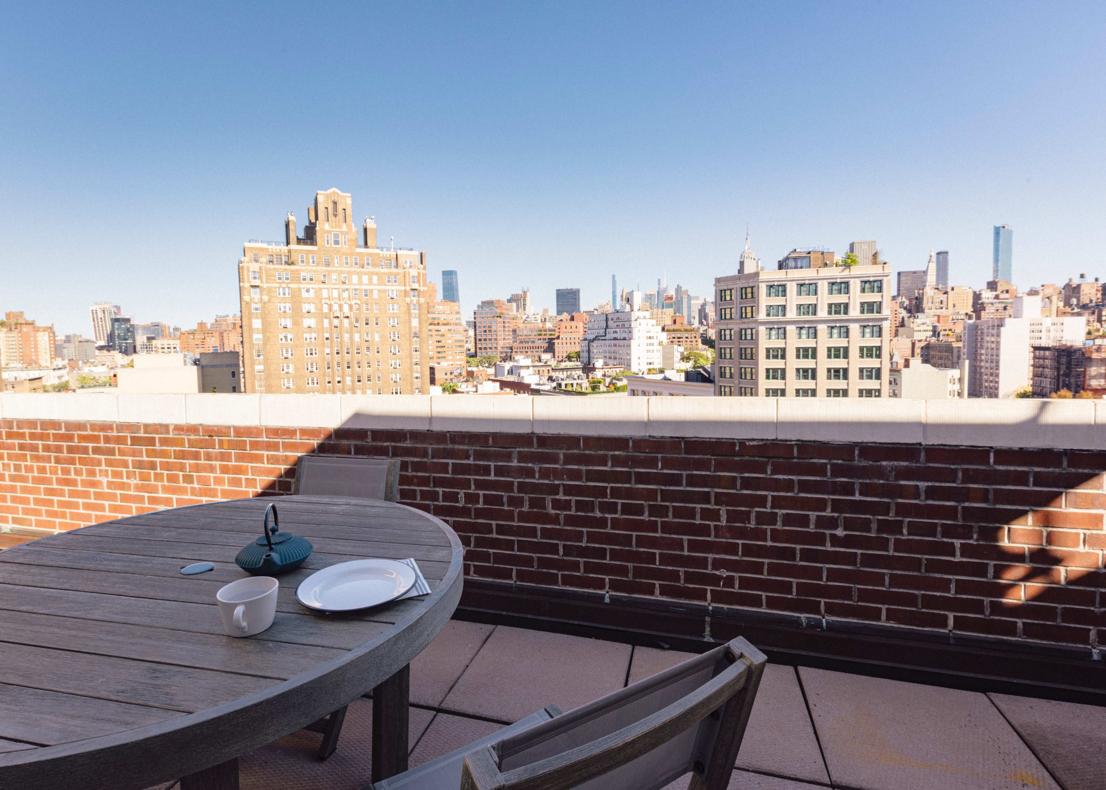 A wooden table with a plate and cup is set on a rooftop terrace overlooking a city skyline.