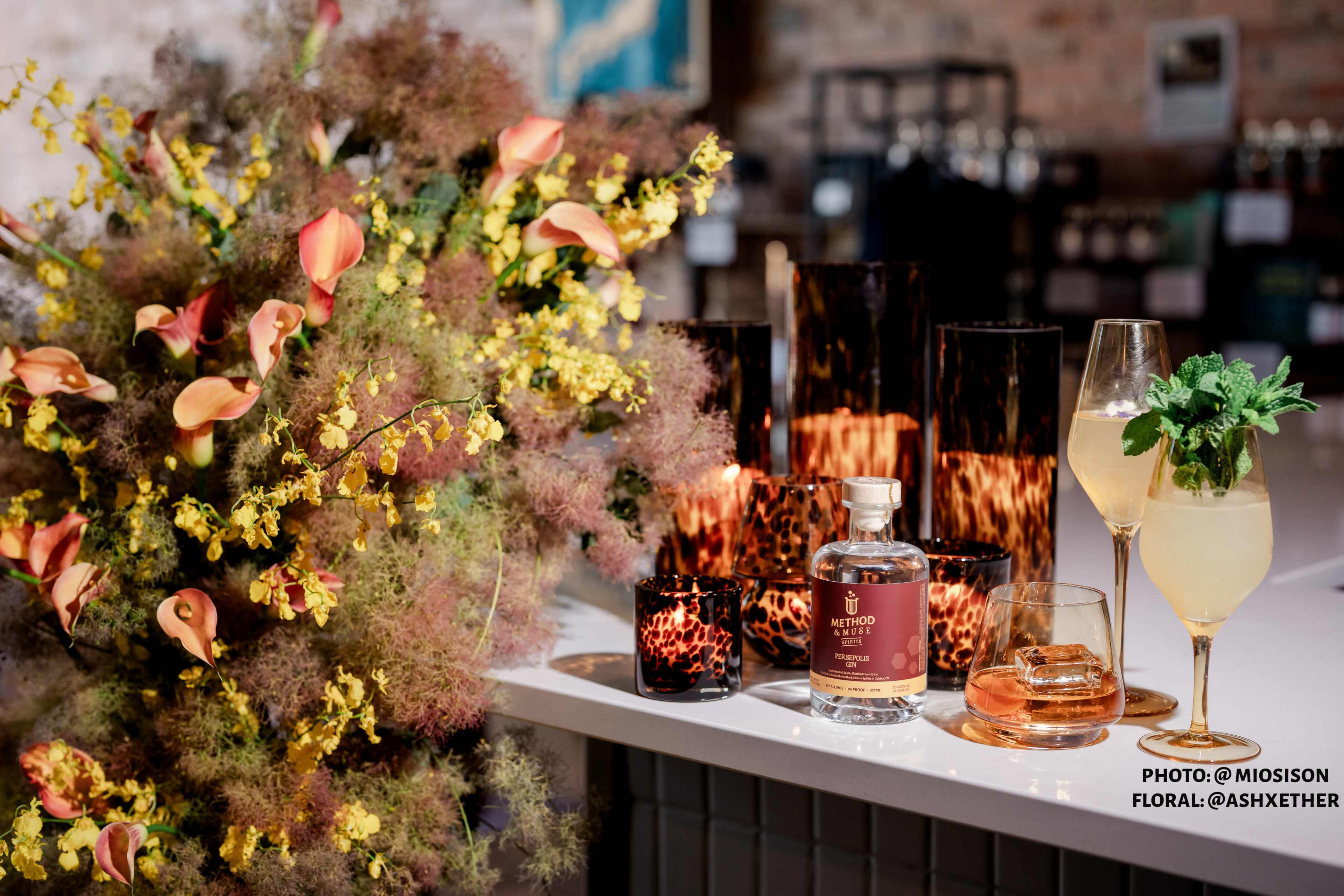 A floral arrangement with yellow and orange flowers is positioned next to a bottle of liquor and two cocktails on a bar counter.