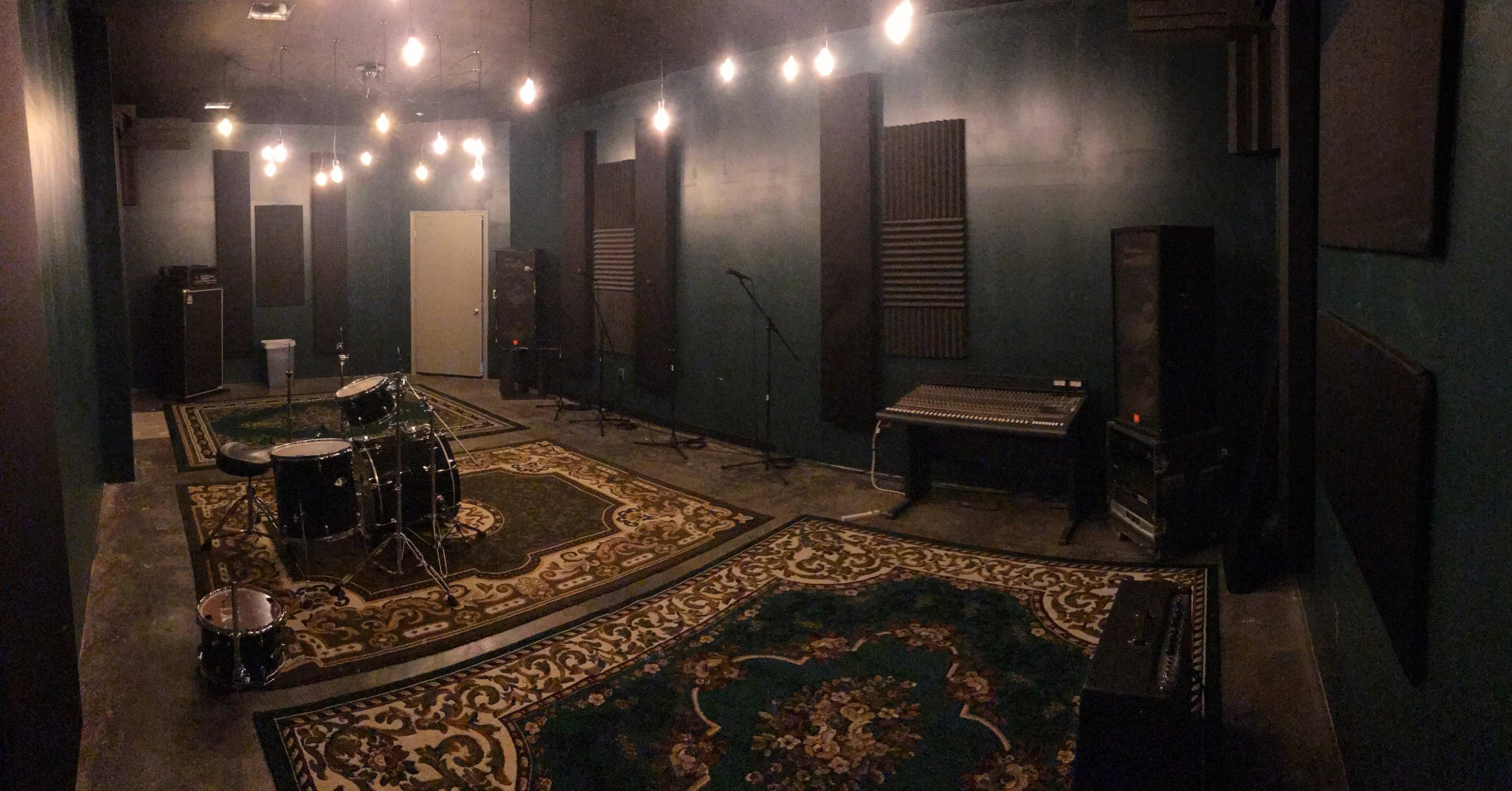 A dimly lit music practice room with a drum set, keyboard, and sound equipment arranged on patterned rugs.