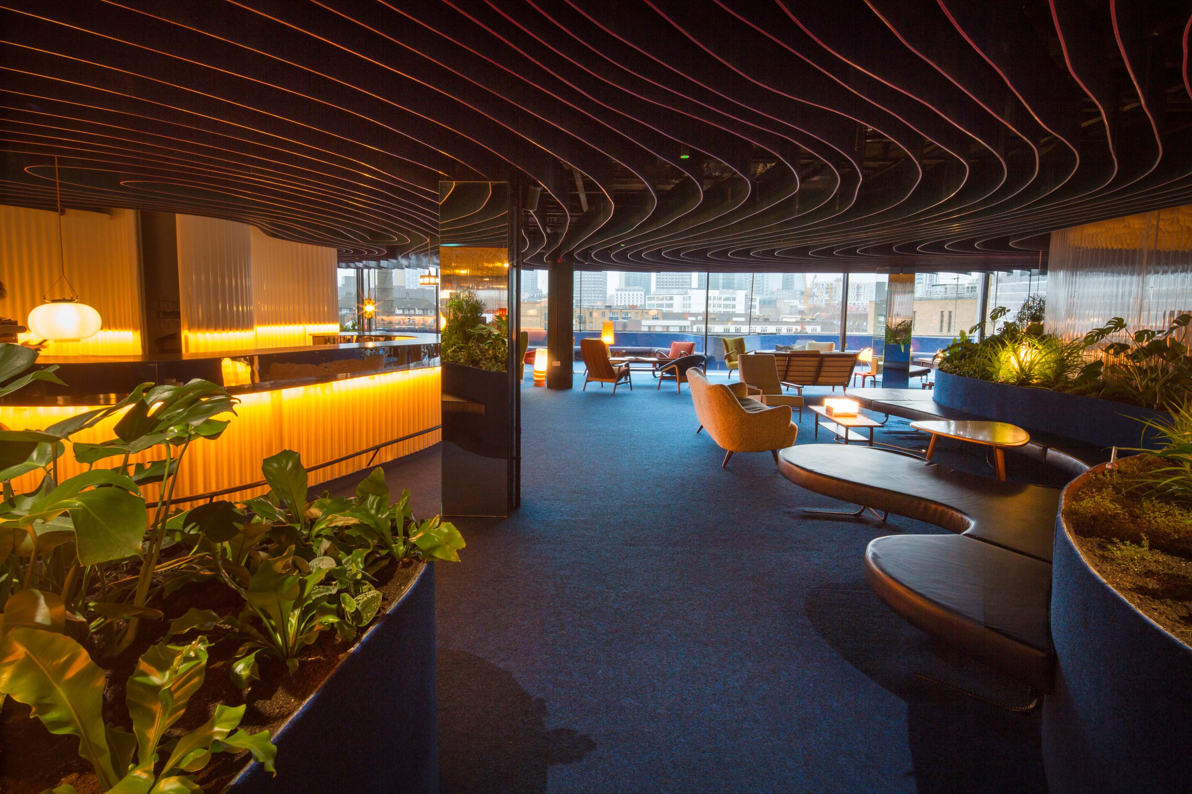 The image shows a modern interior of a lounge area featuring curved walls, plants, and various seating arrangements with large windows overlooking a cityscape.