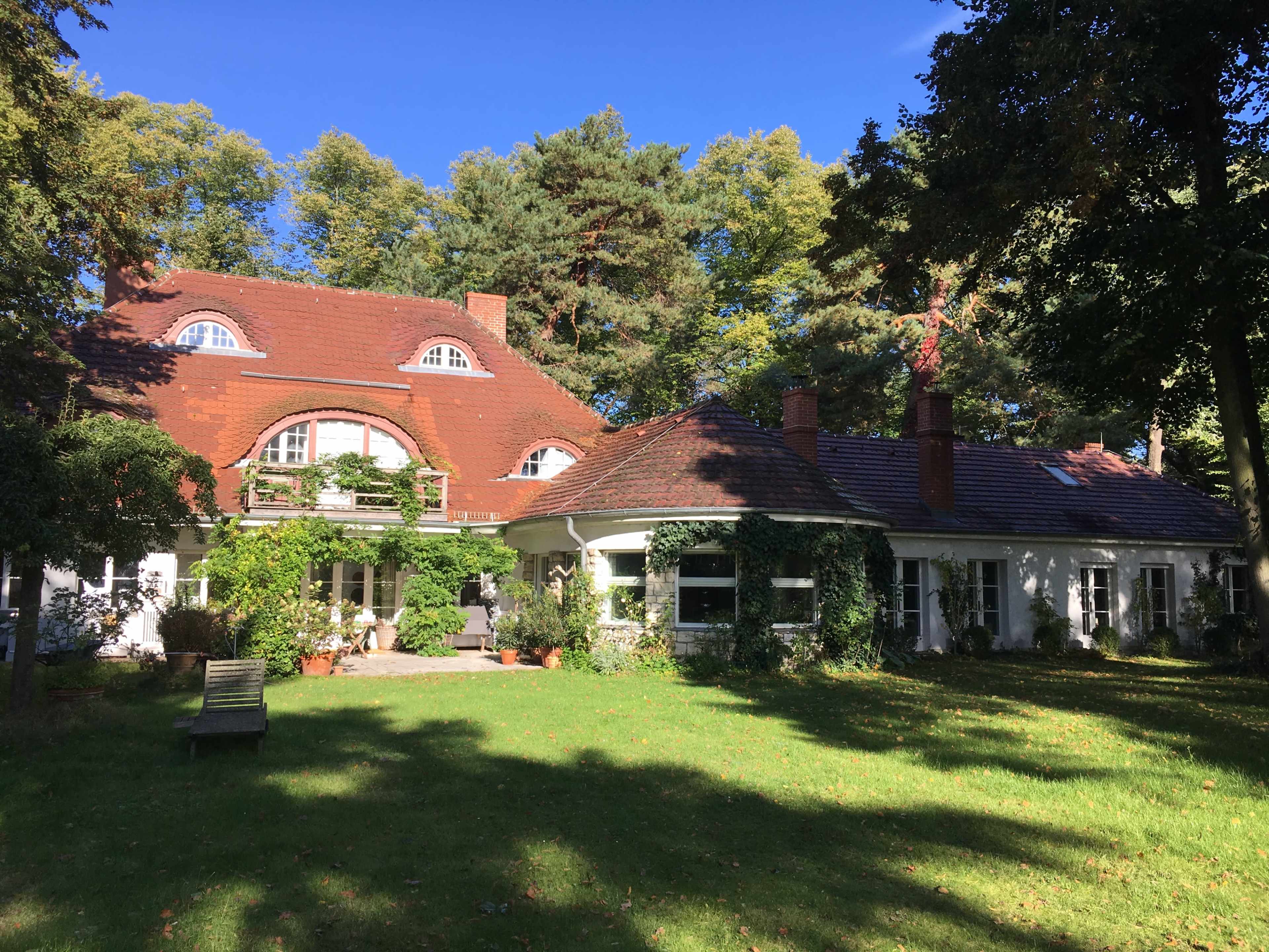 A large, two-story house with a red tiled roof is surrounded by trees and green grass.