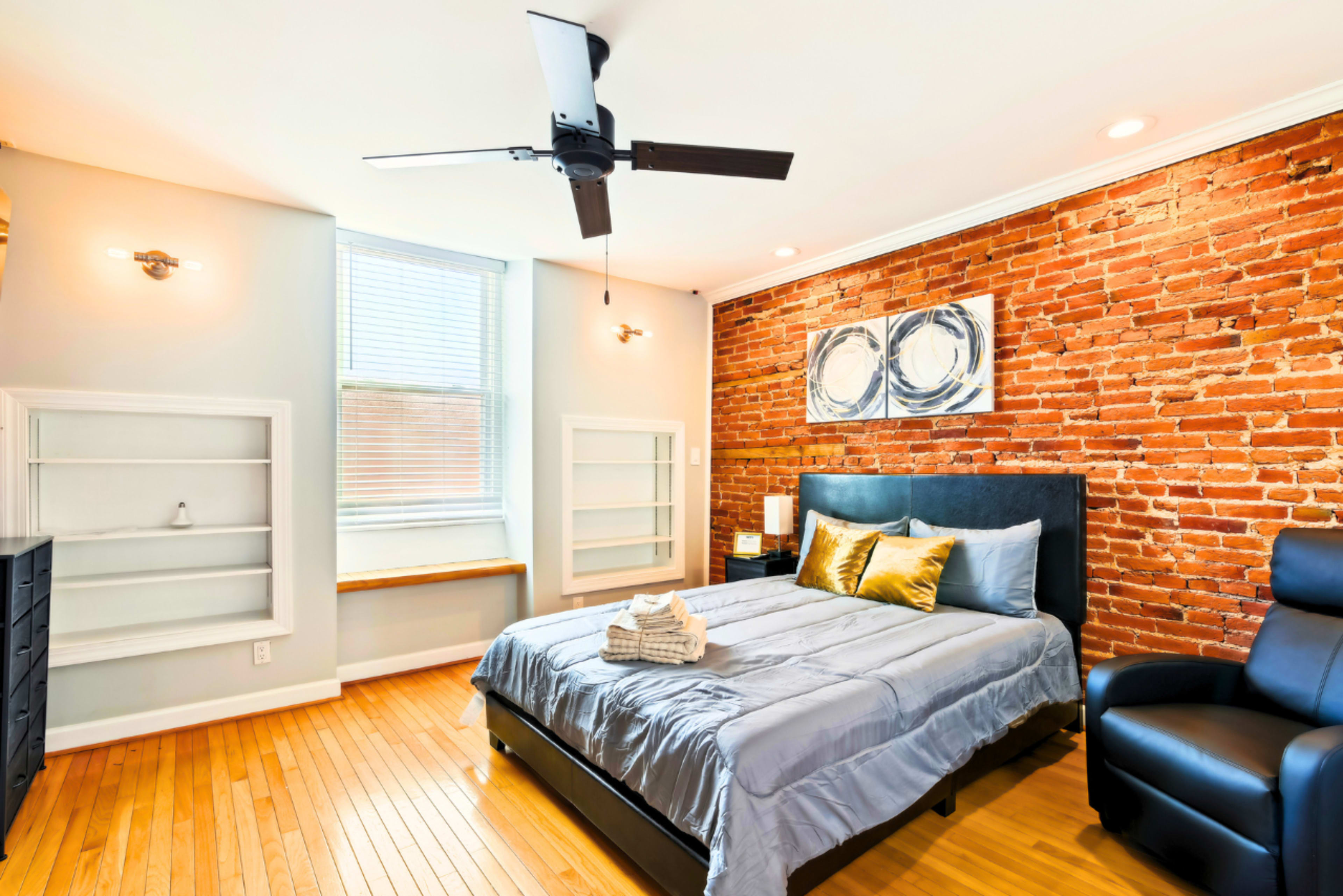 The image shows a modern bedroom featuring a large bed with gray bedding, an exposed brick wall, a wooden floor, a ceiling fan, and a window with natural light.