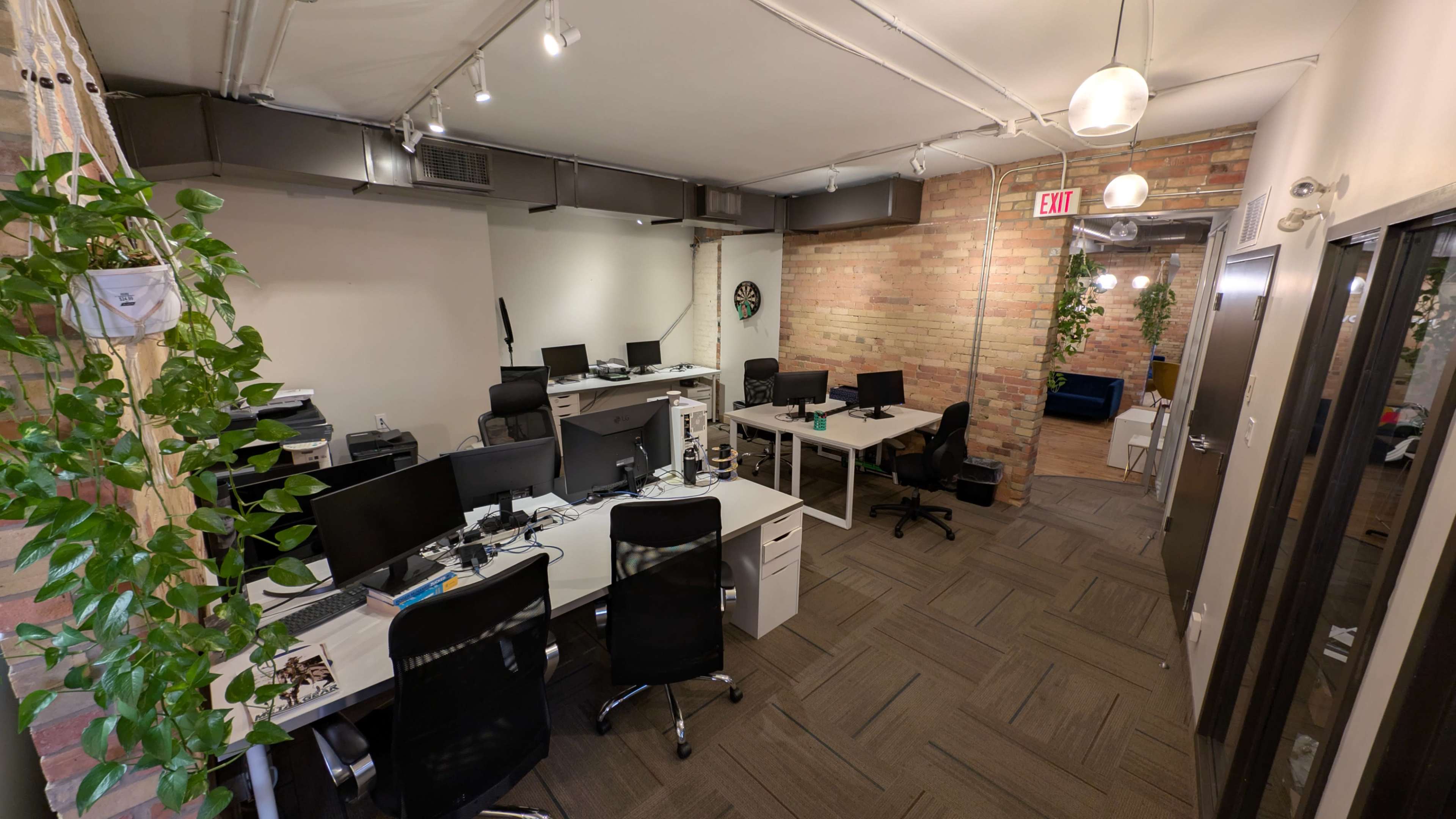 The image shows a modern office space with multiple desks equipped with computers, potted plants, and exposed brick walls.