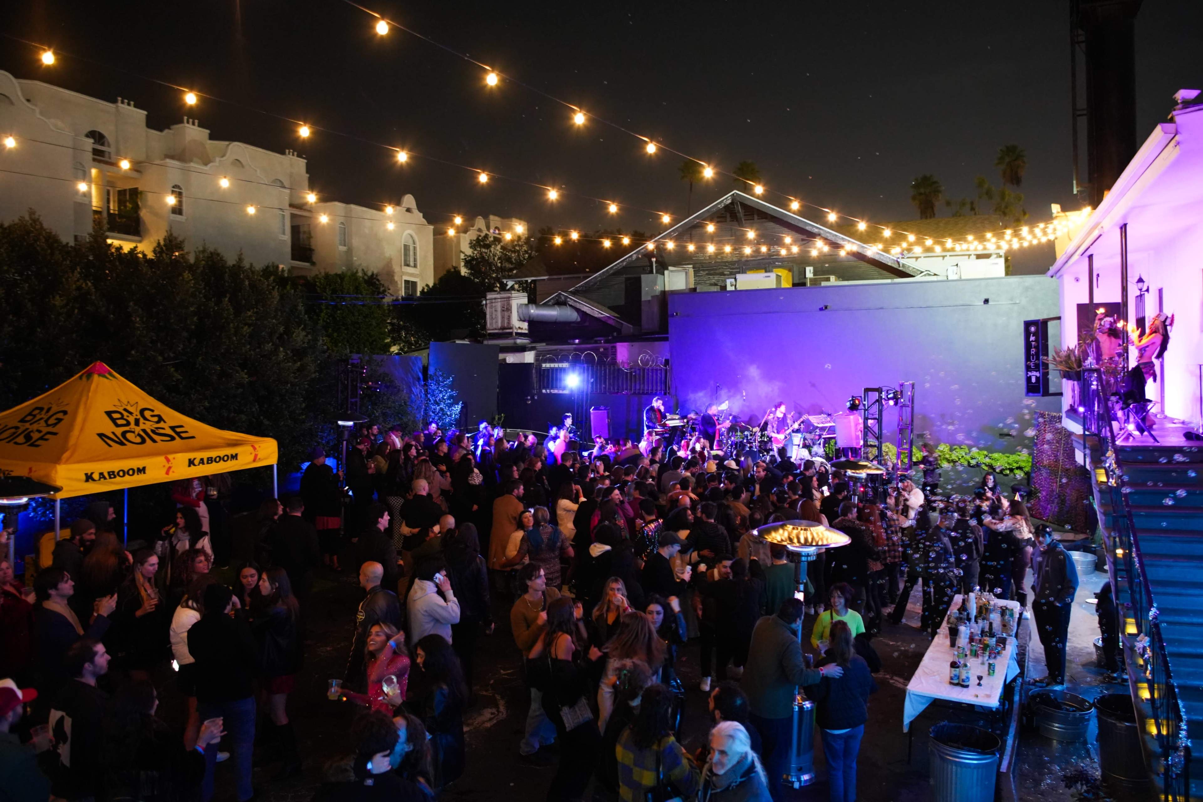 A large crowd gathers at a nighttime outdoor event with string lights overhead, a stage for live music, and a bar area.