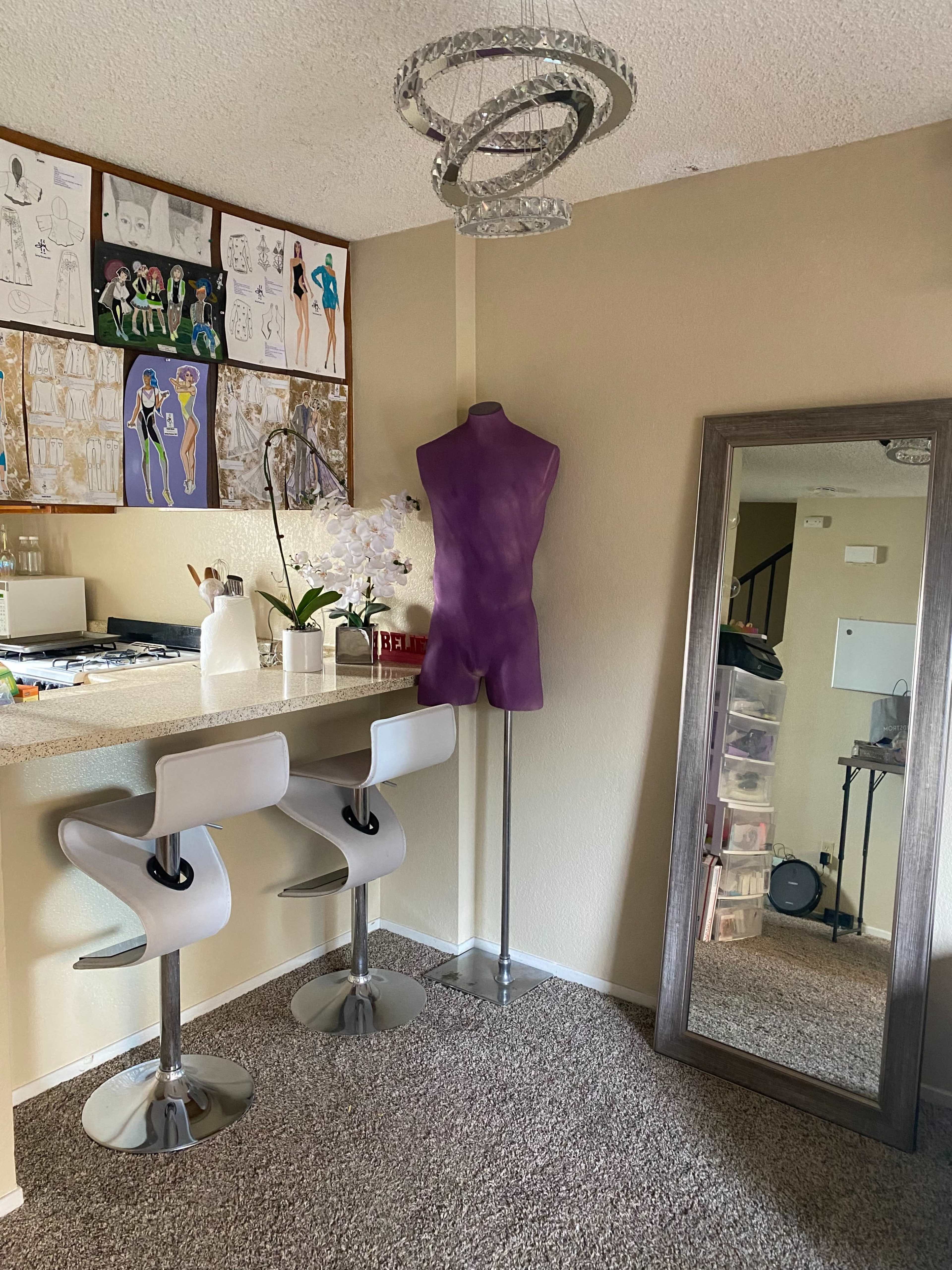 A room with a purple dress form beside a large mirror and two modern bar stools at a countertop, with fashion sketches displayed on the wall.