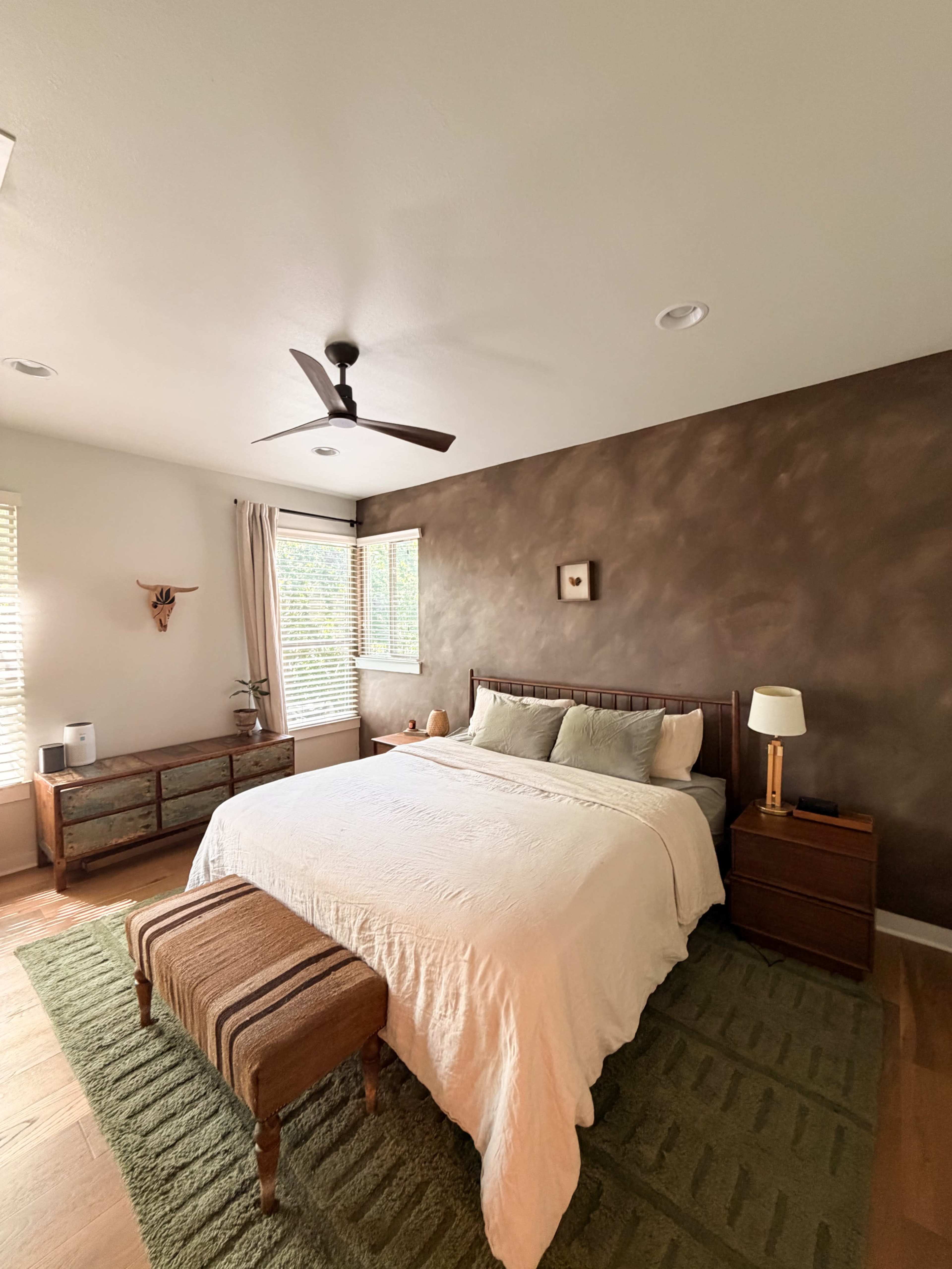 The image shows a simple bedroom with a large bed, a wooden bench at the foot, a nightstand, and a dresser under windows with natural light.