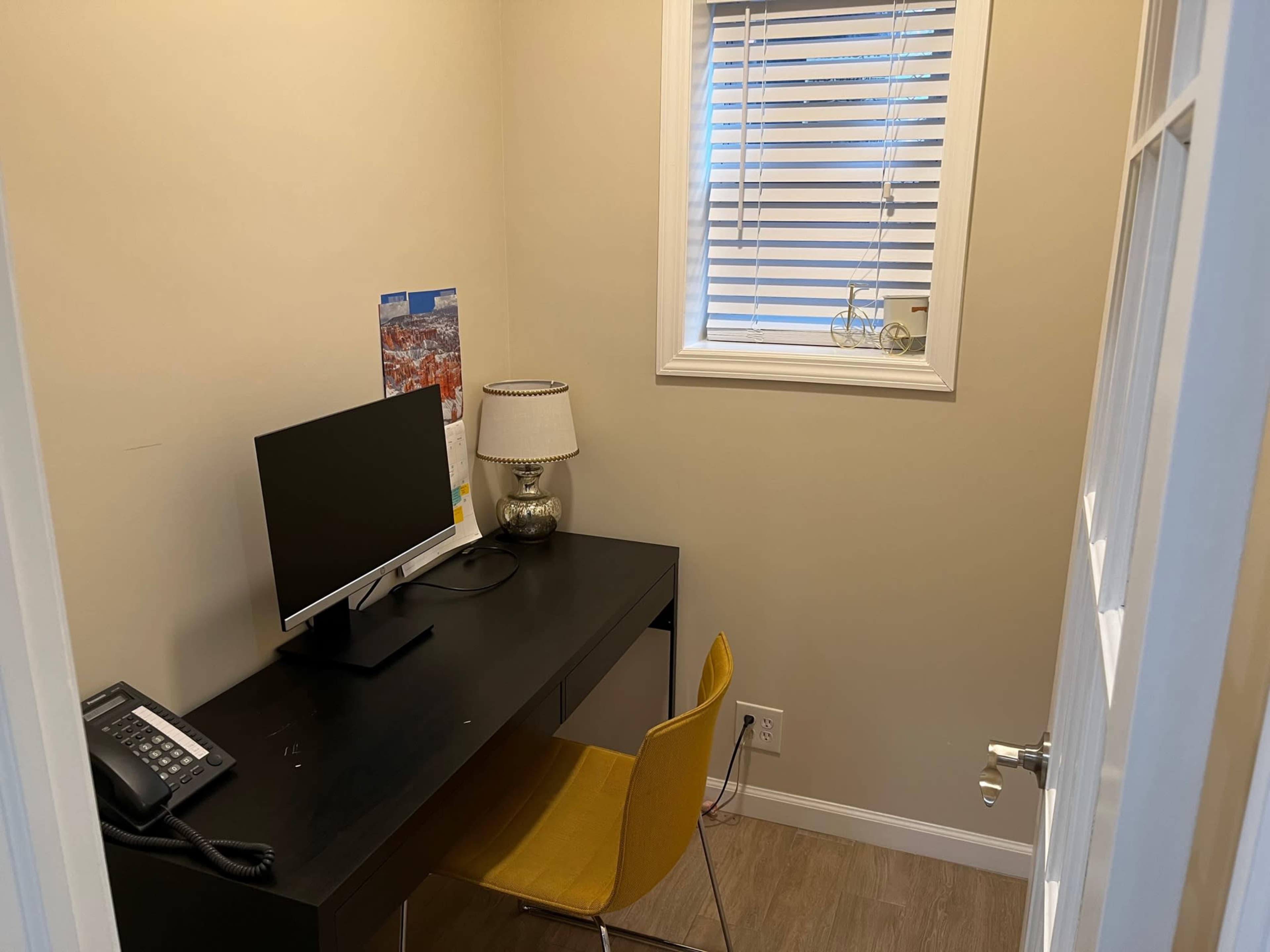 A small office space features a black desk with a monitor, a telephone, a lamp, and a yellow chair, with a window allowing natural light.