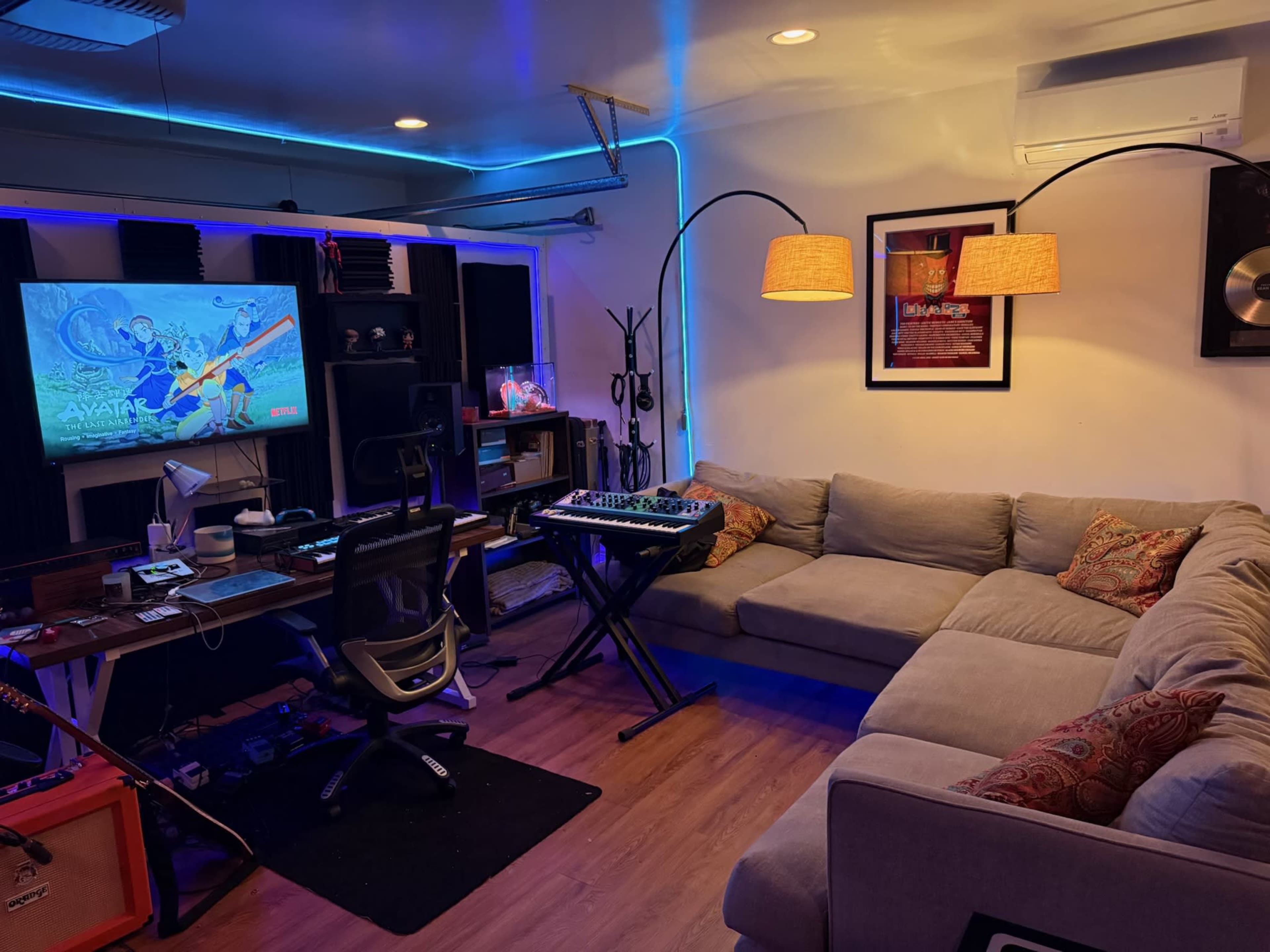 The image shows a cozy studio space featuring a large sectional couch, an electronic keyboard on a stand, and a television displaying promotional material for "Avatar."