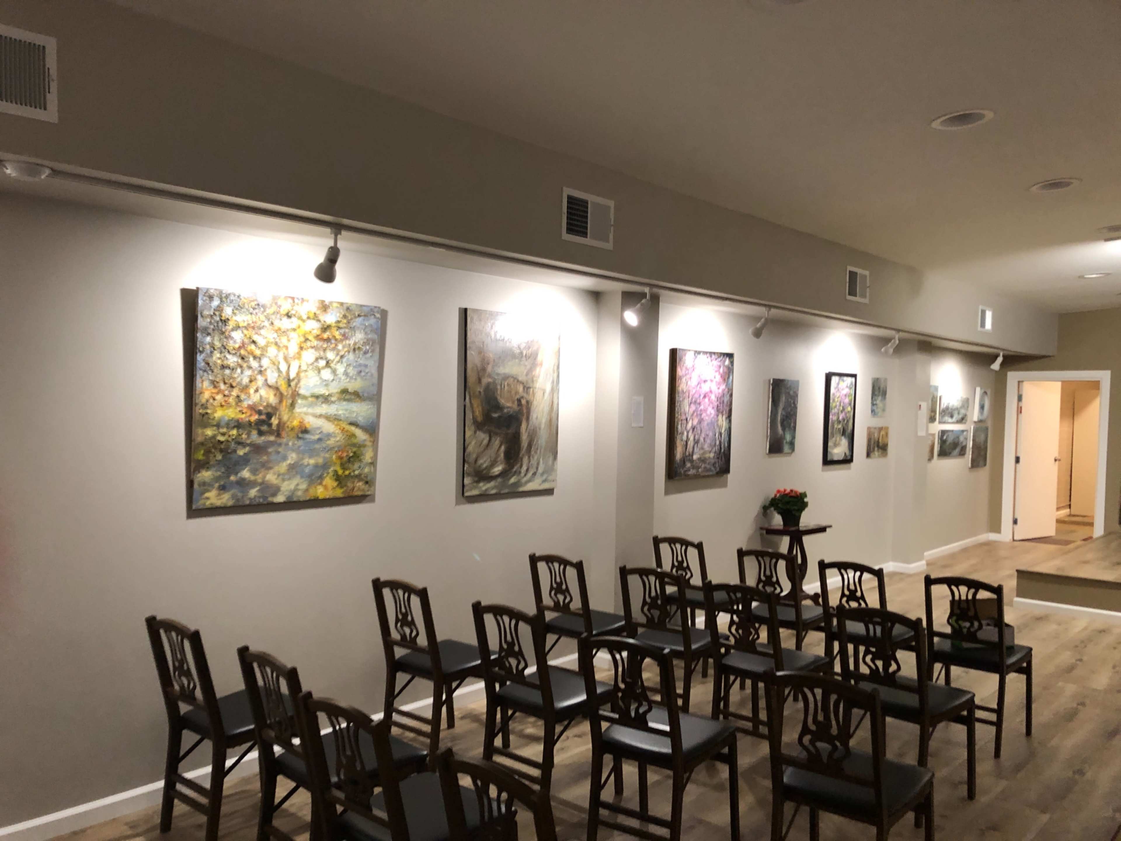 A gallery with a row of paintings on the wall and a series of empty chairs arranged in front of them.