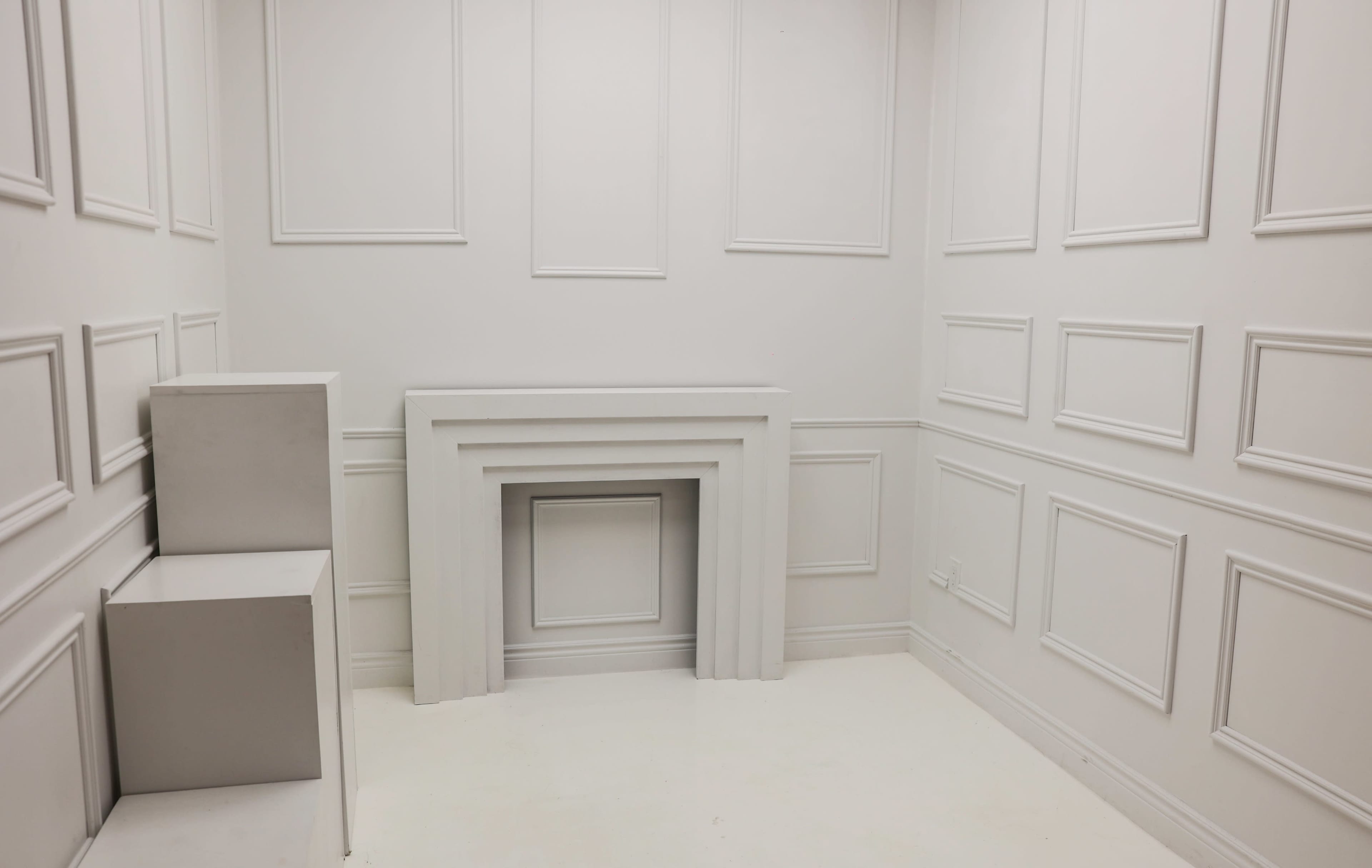 The image shows a minimalist white room with paneled walls and a simple, decorative fireplace.