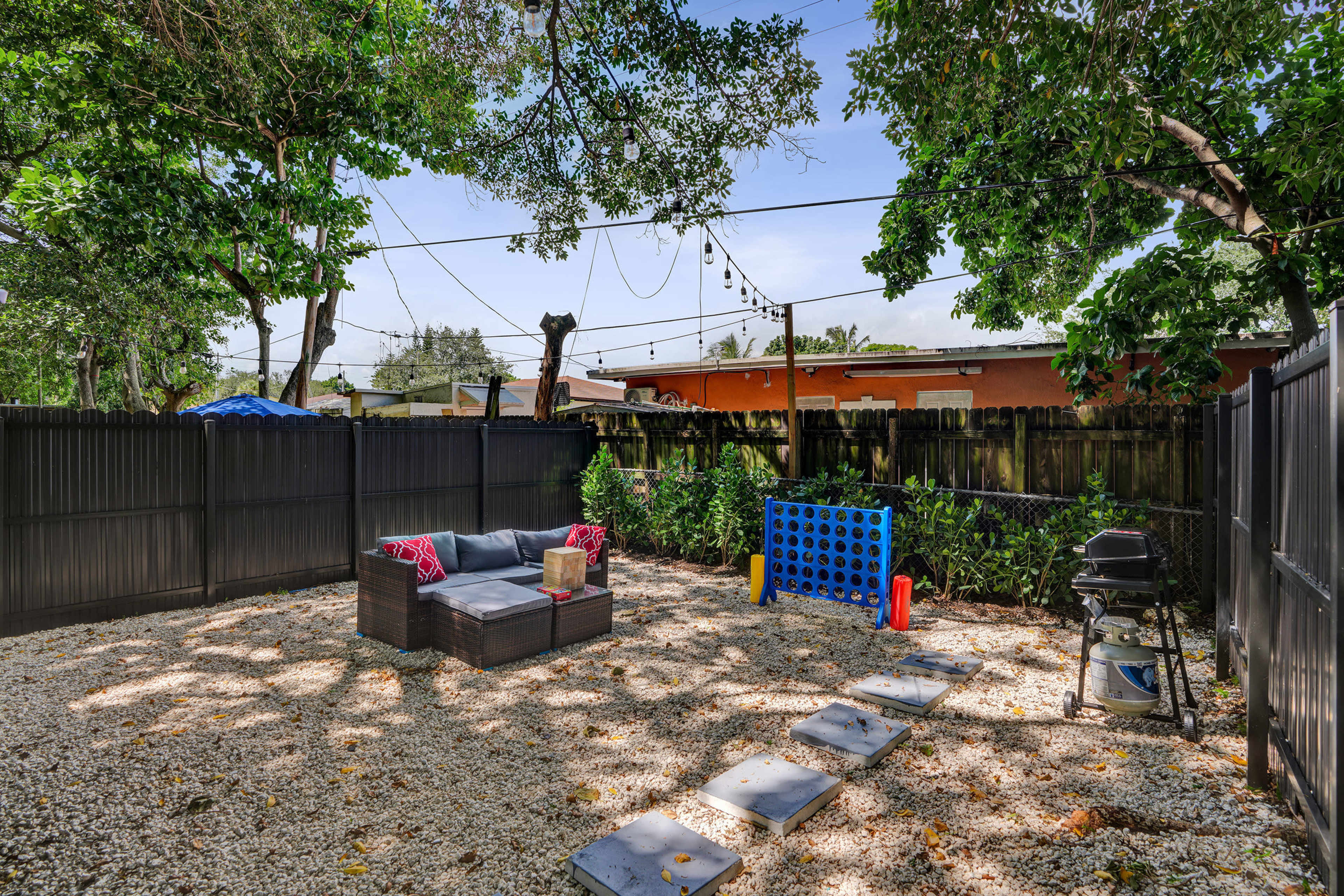 The image shows a gravel backyard with a seating area, a large blue Connect Four game, and a barbecue grill, surrounded by trees and a black fence.
