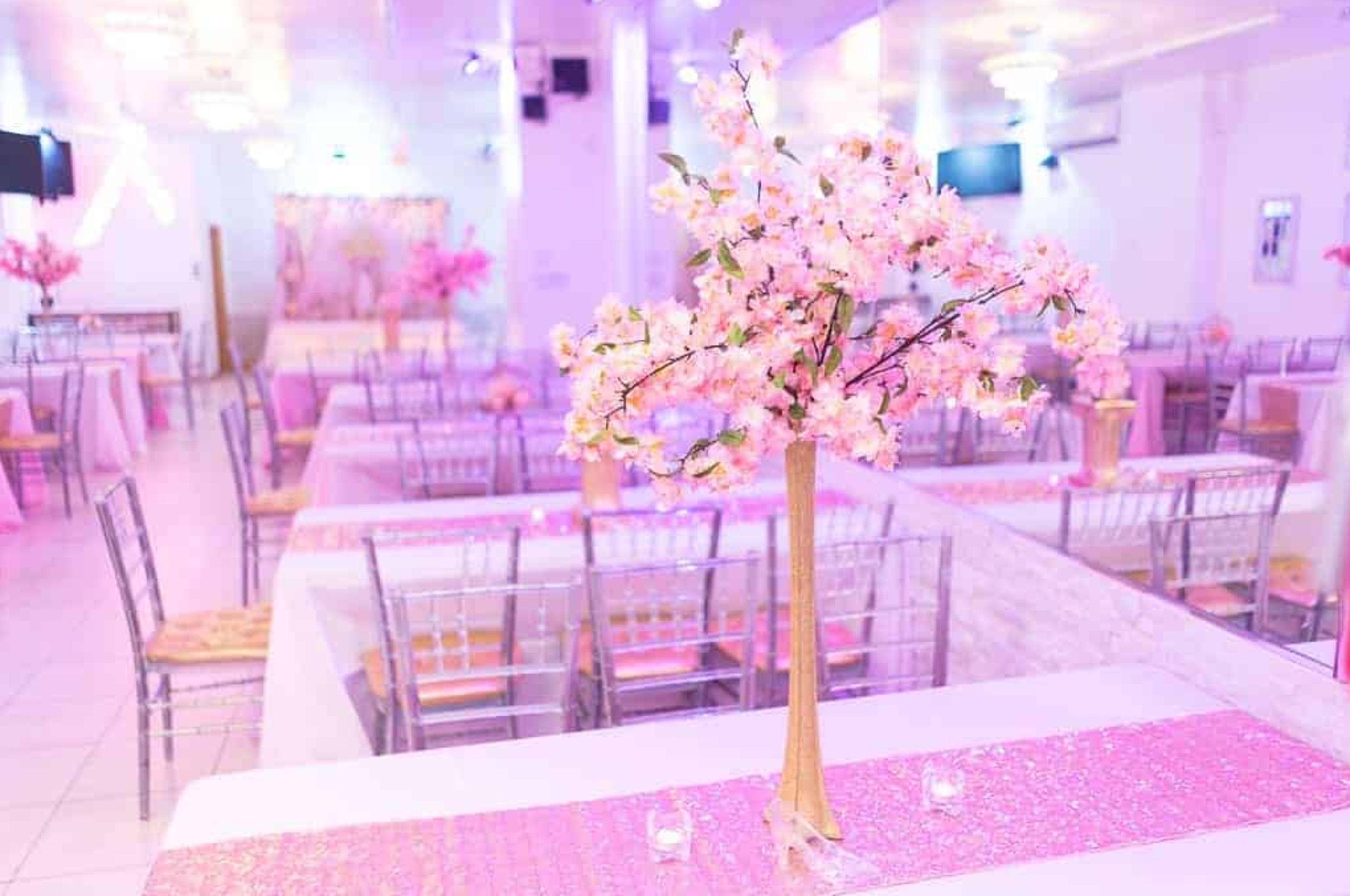 The image shows a banquet hall decorated with pink floral arrangements and tables set with elegant tablecloths and centerpieces.