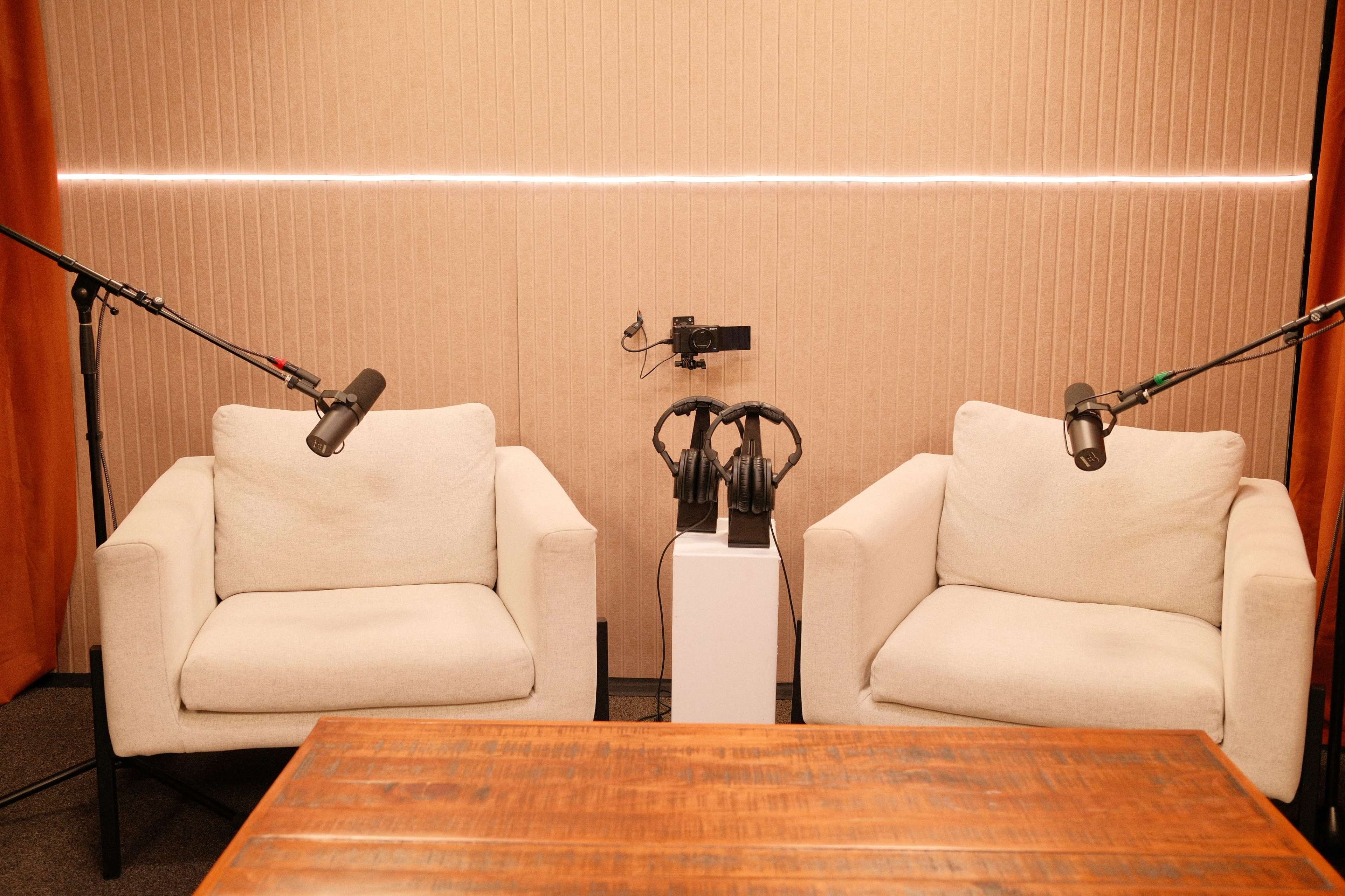 The image shows two white armchairs positioned in front of a wooden table, with microphones and headphones arranged on a stand between them in a soundproof studio.