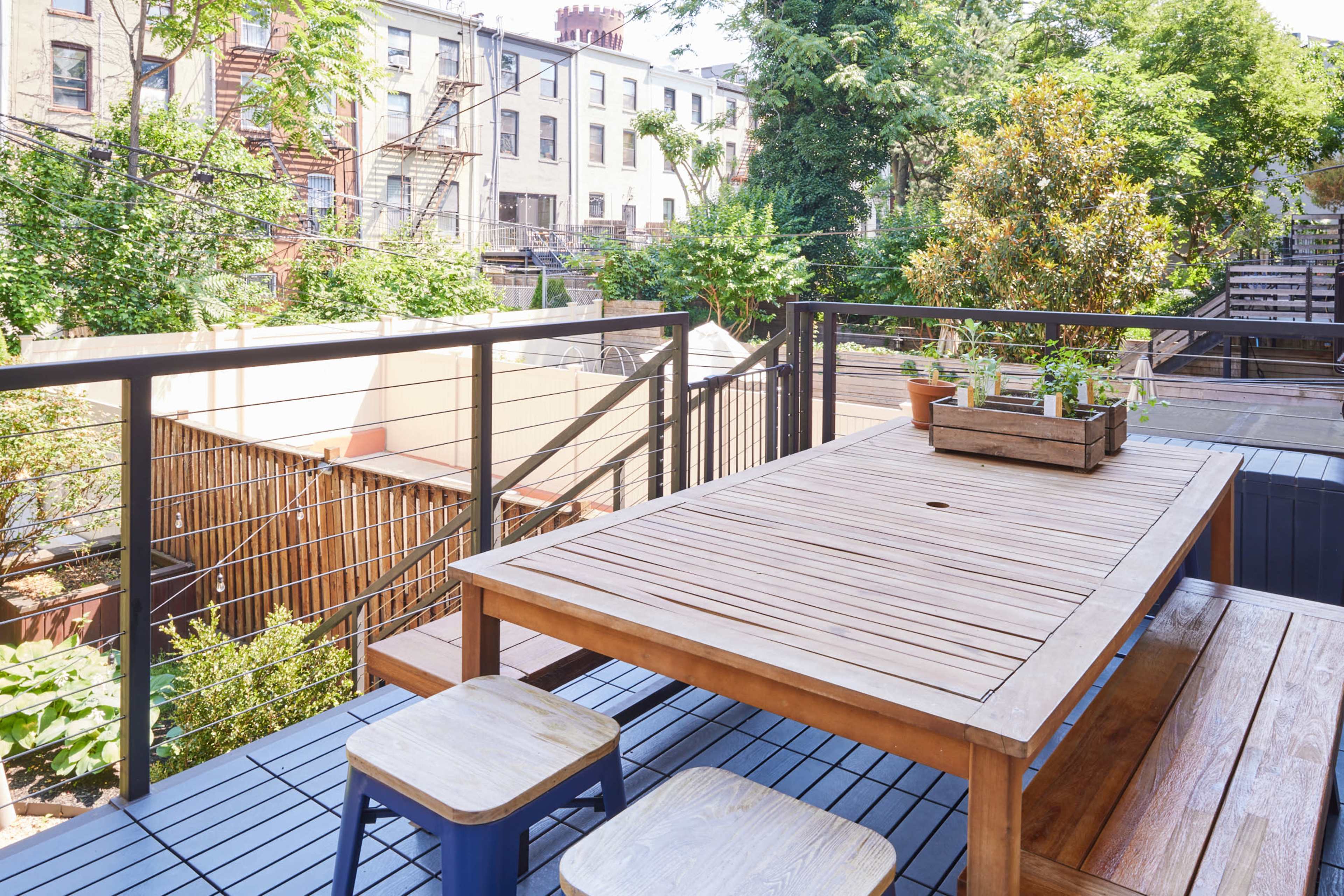 A wooden dining table and two stools are positioned on a deck overlooking a green backyard with trees and nearby buildings.