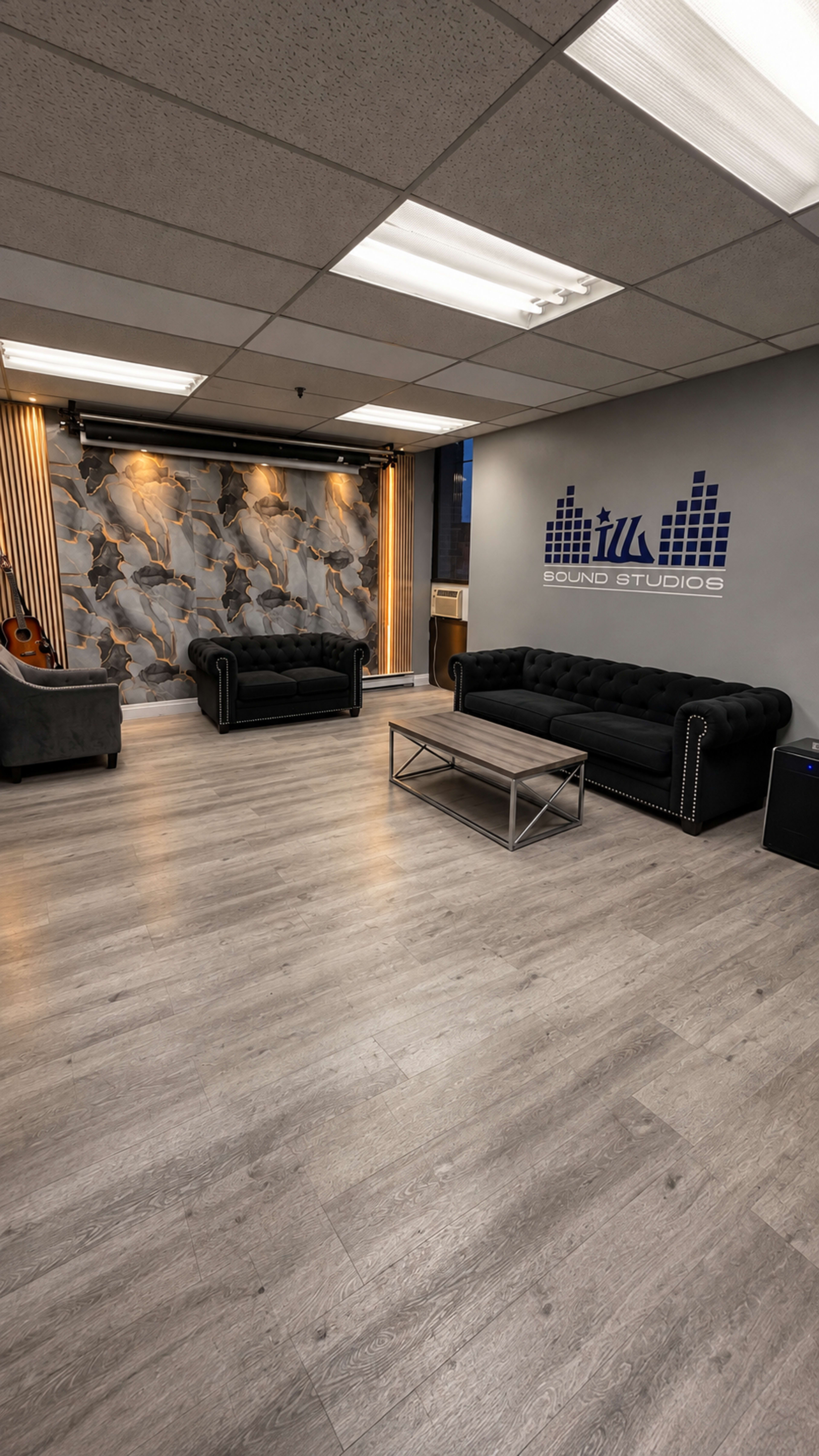 The image shows a modern sound studio featuring two black sofas, a coffee table, and a textured wall design.