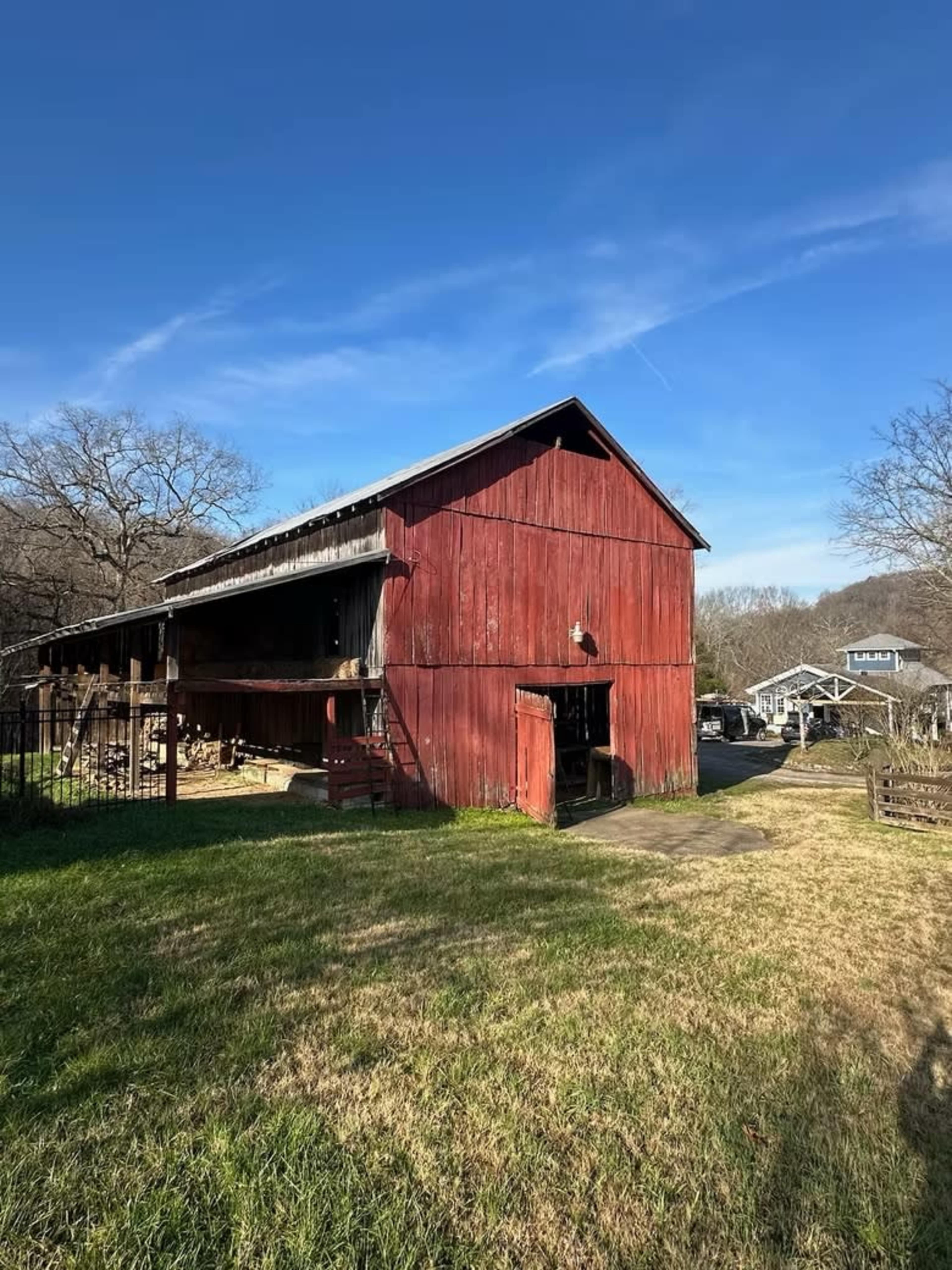 A weathered red barn stands beside a grassy area, with a clear blue sky above and homes visible in the background.