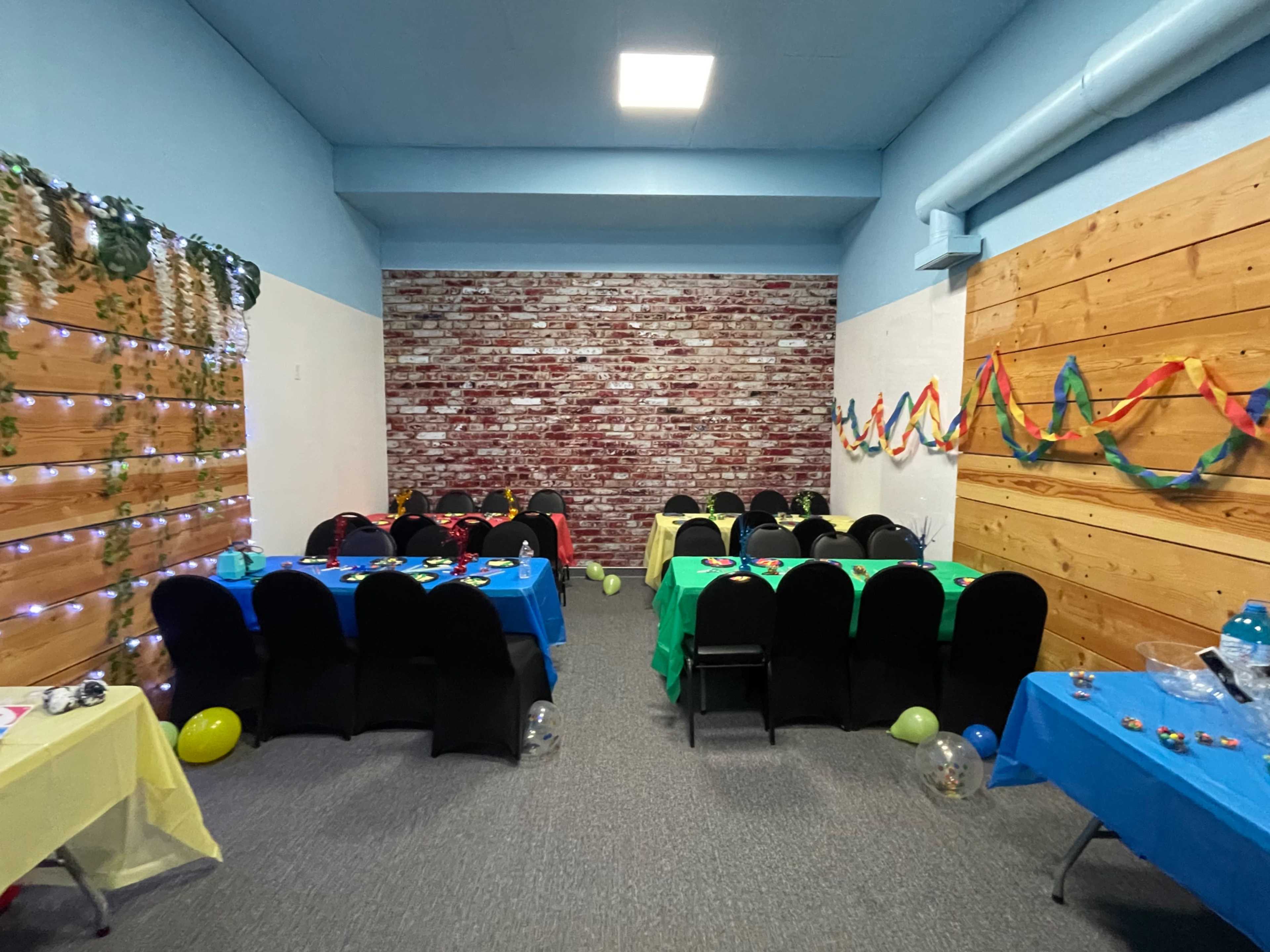 The image shows a decorated room with six tables covered in colorful tablecloths, surrounded by balloons and streamers, against a backdrop of a brick and wooden wall.