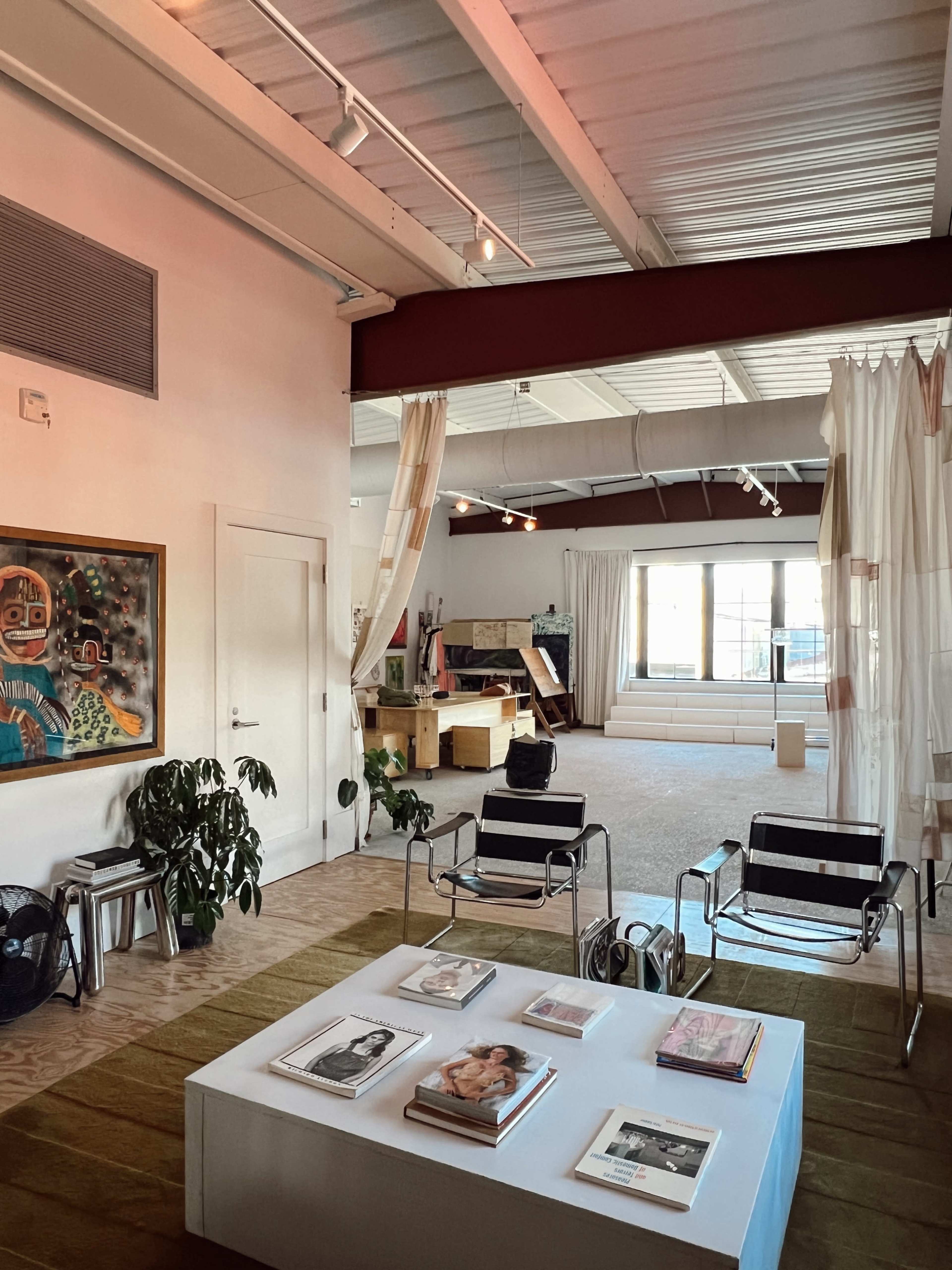 The image features a spacious, well-lit studio with modern furniture, large windows, and a collection of magazines displayed on a coffee table.