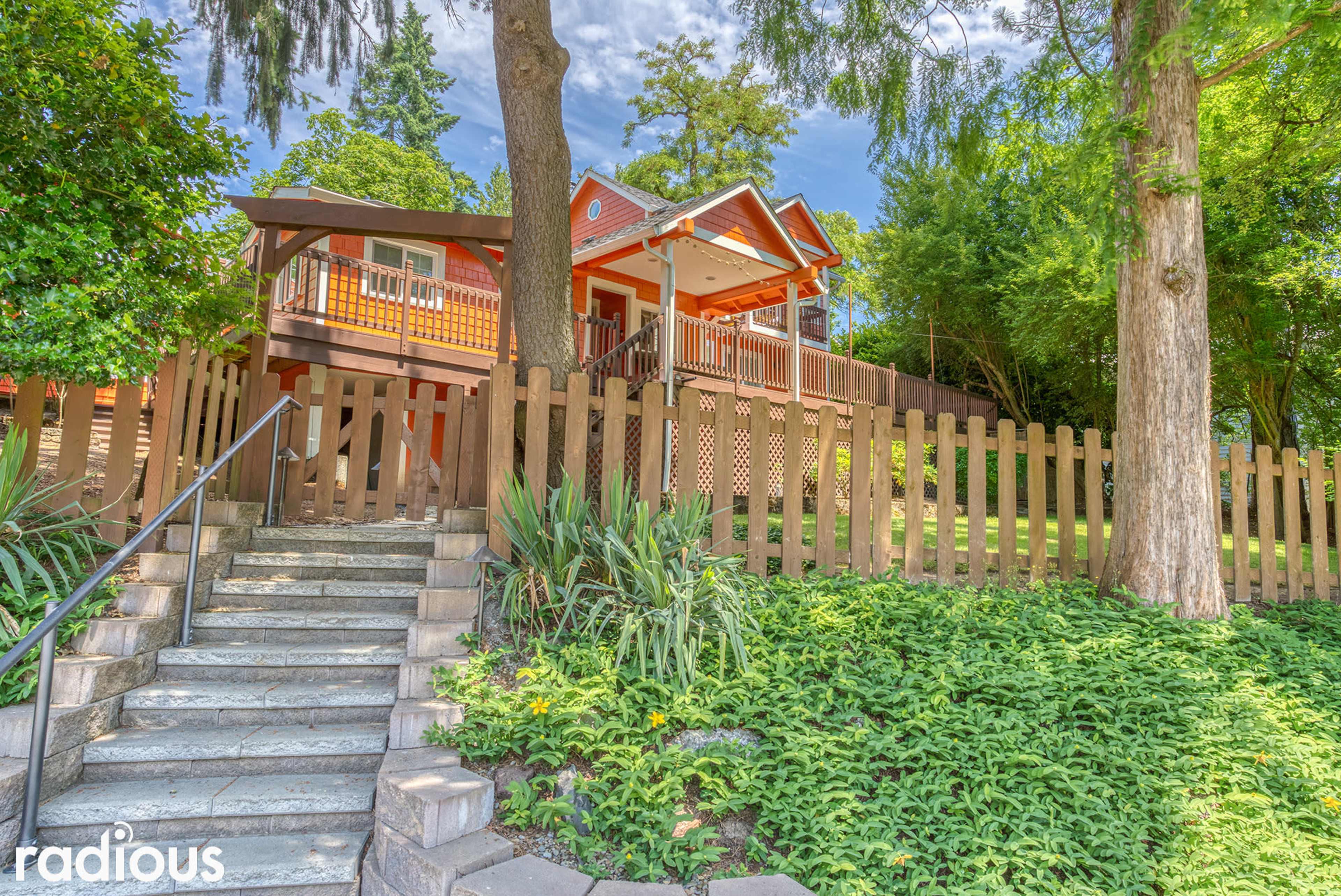 A brightly colored house with a porch and wooden railing is set behind a low fence, surrounded by greenery and trees.