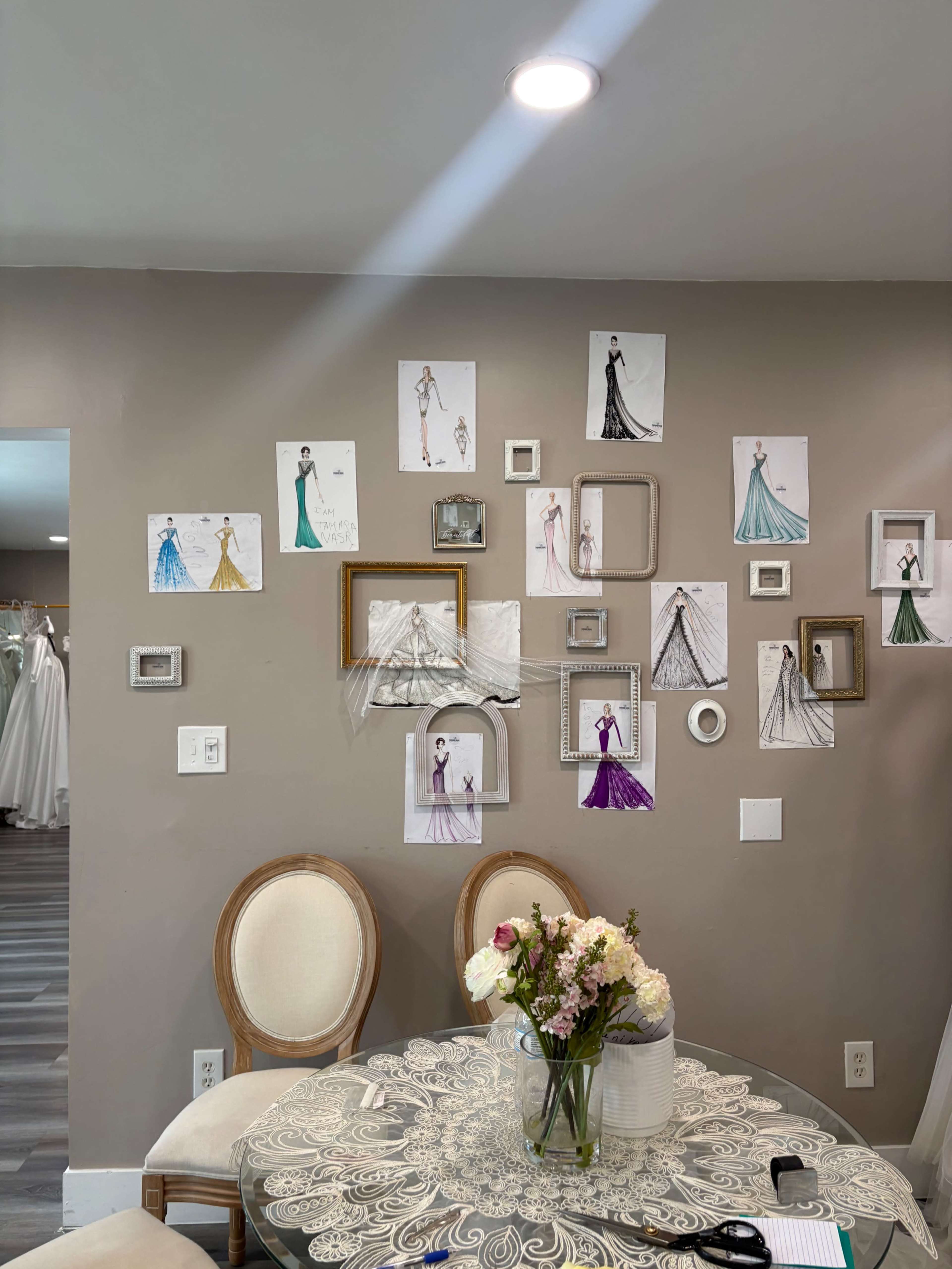 A wall adorned with fashion illustrations and framed sketches of dresses is complemented by a circular table with a vase of flowers in the center.